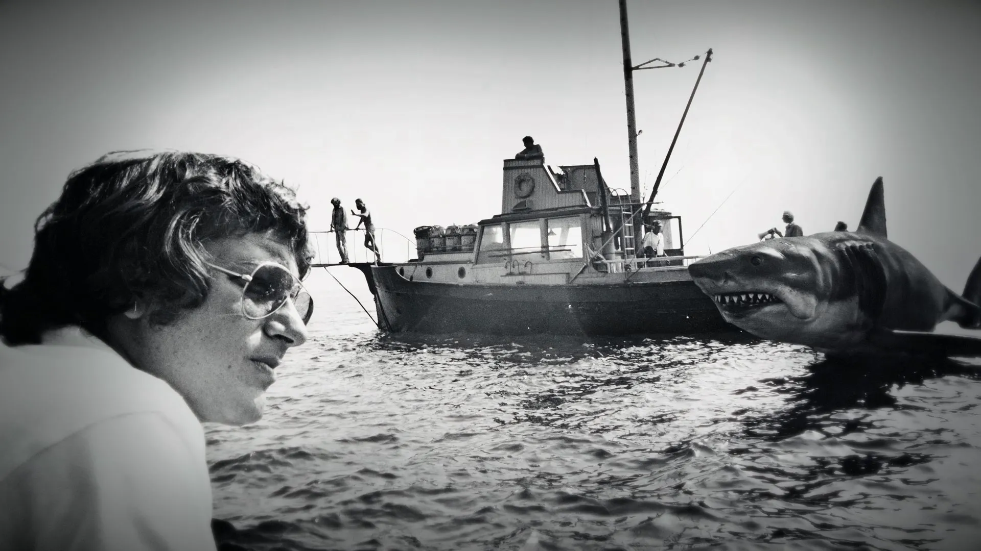 Black and white behind-the-scenes shot of a young Steven Spielberg on the set of Jaws with the boat and iconic shark model in the background.