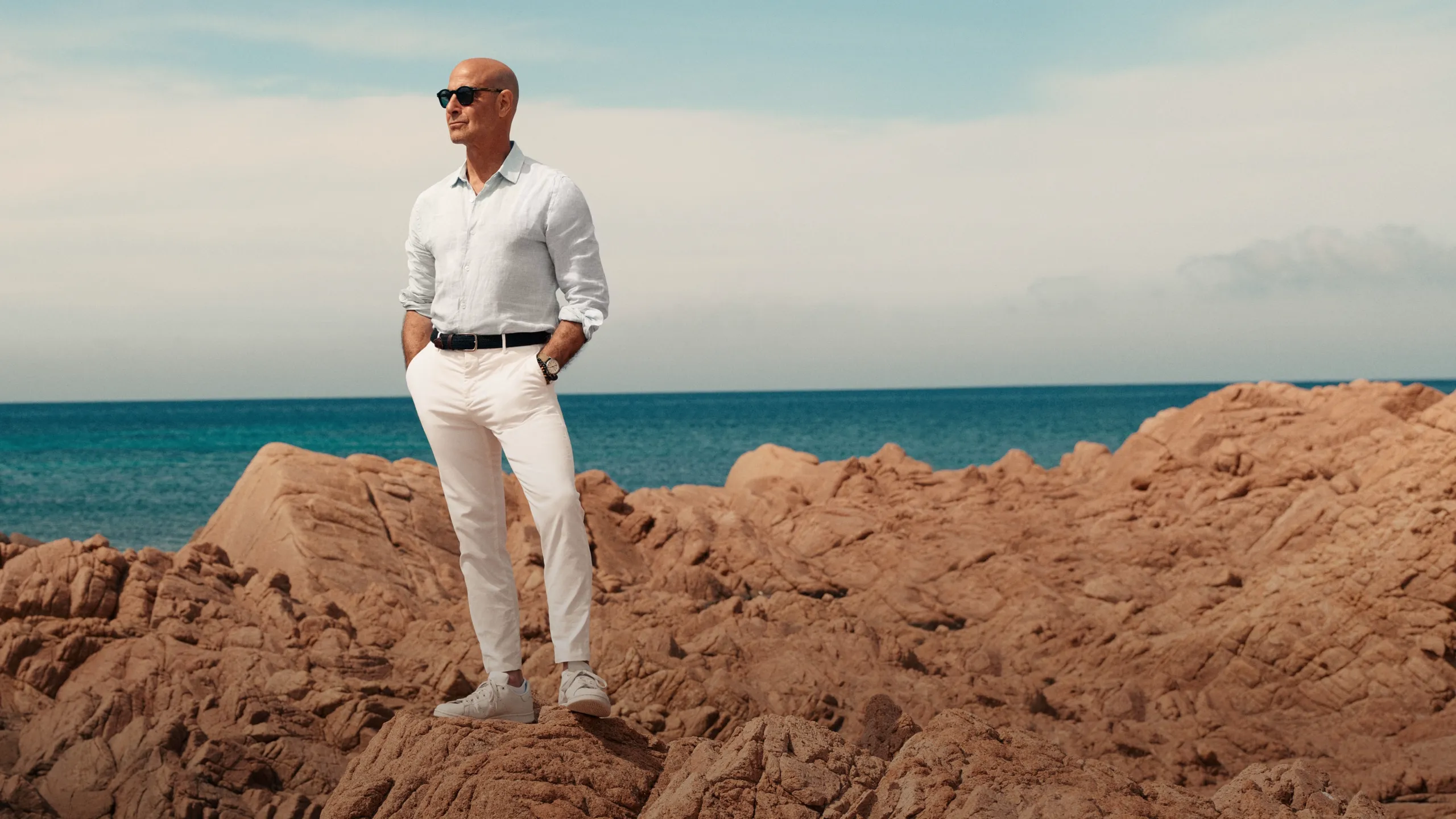 Stanley Tucci stands on some cliffs in Italy, dressed in white and wearing sunglasses