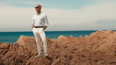 Stanley Tucci stands on some cliffs in Italy, dressed in white and wearing sunglasses
