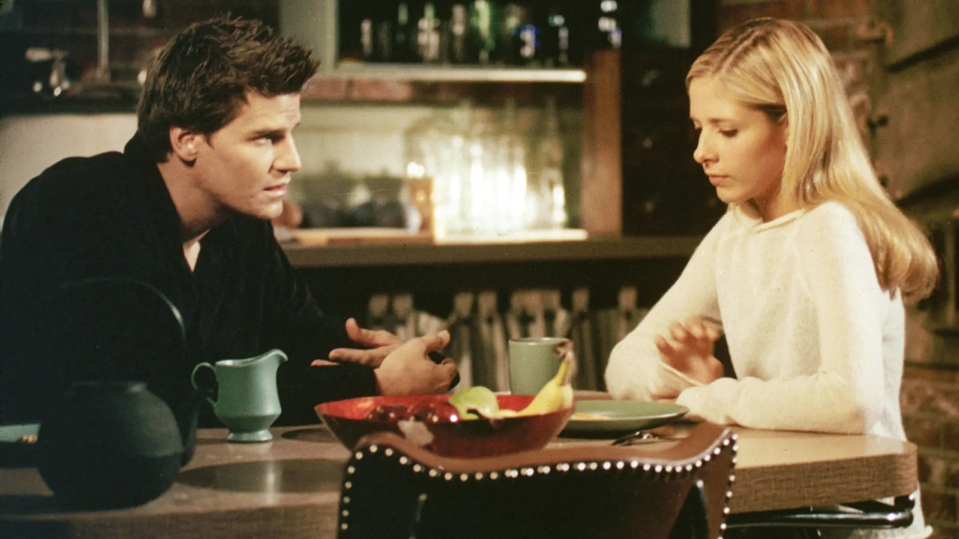 Buffy Summers (Sarah Michelle Gellar) sits at a kitchen table to talk with Angel (David Boreanaz) in the crossover episode of Angel, "I Will Remember You".