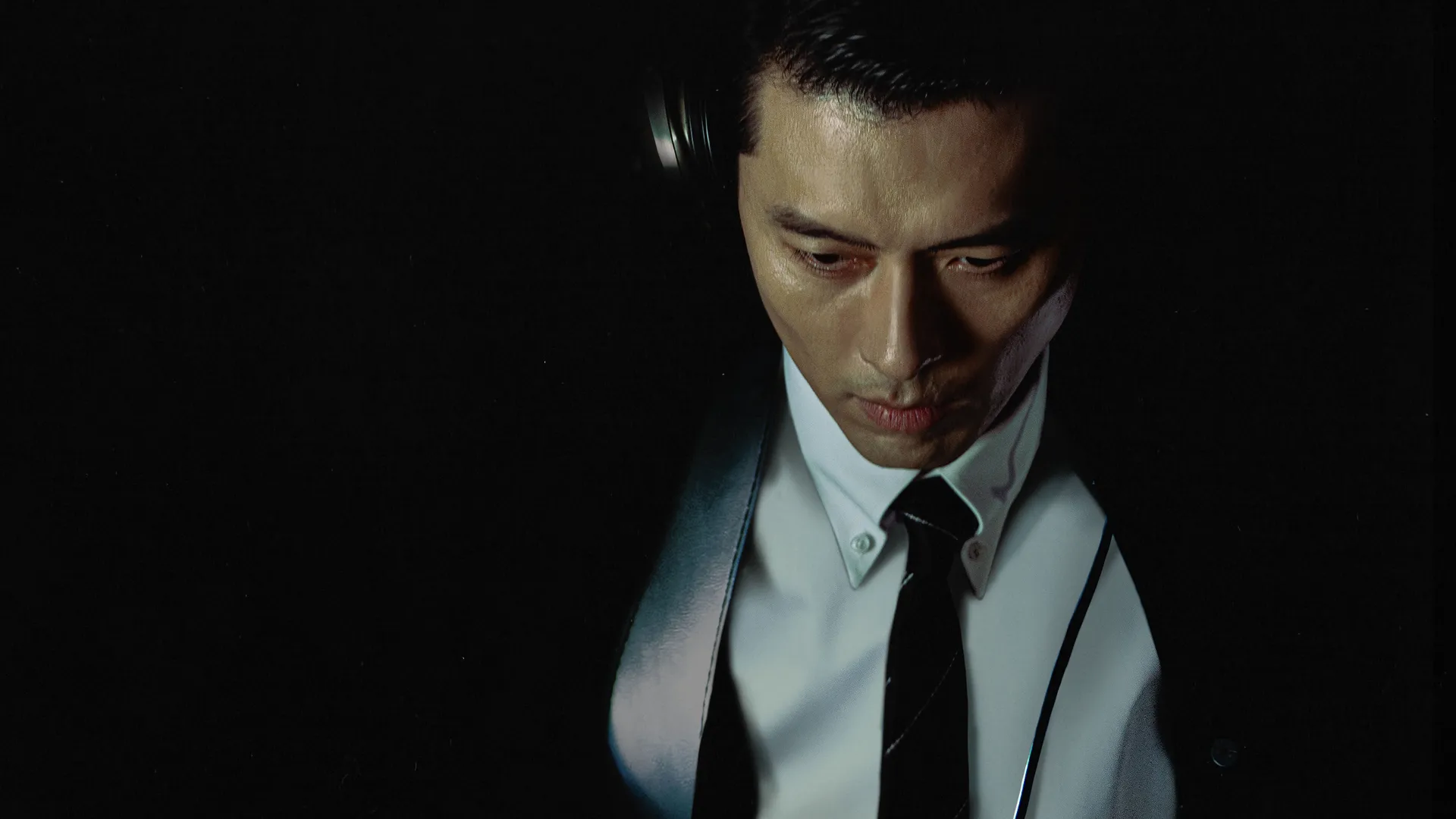 Hyun Bin as the dangerous hostage-taker Min Tae-gu looks down intently while wearing headphones, a white shirt, and a dark tie in a scene from the action thriller film The Negotiation.