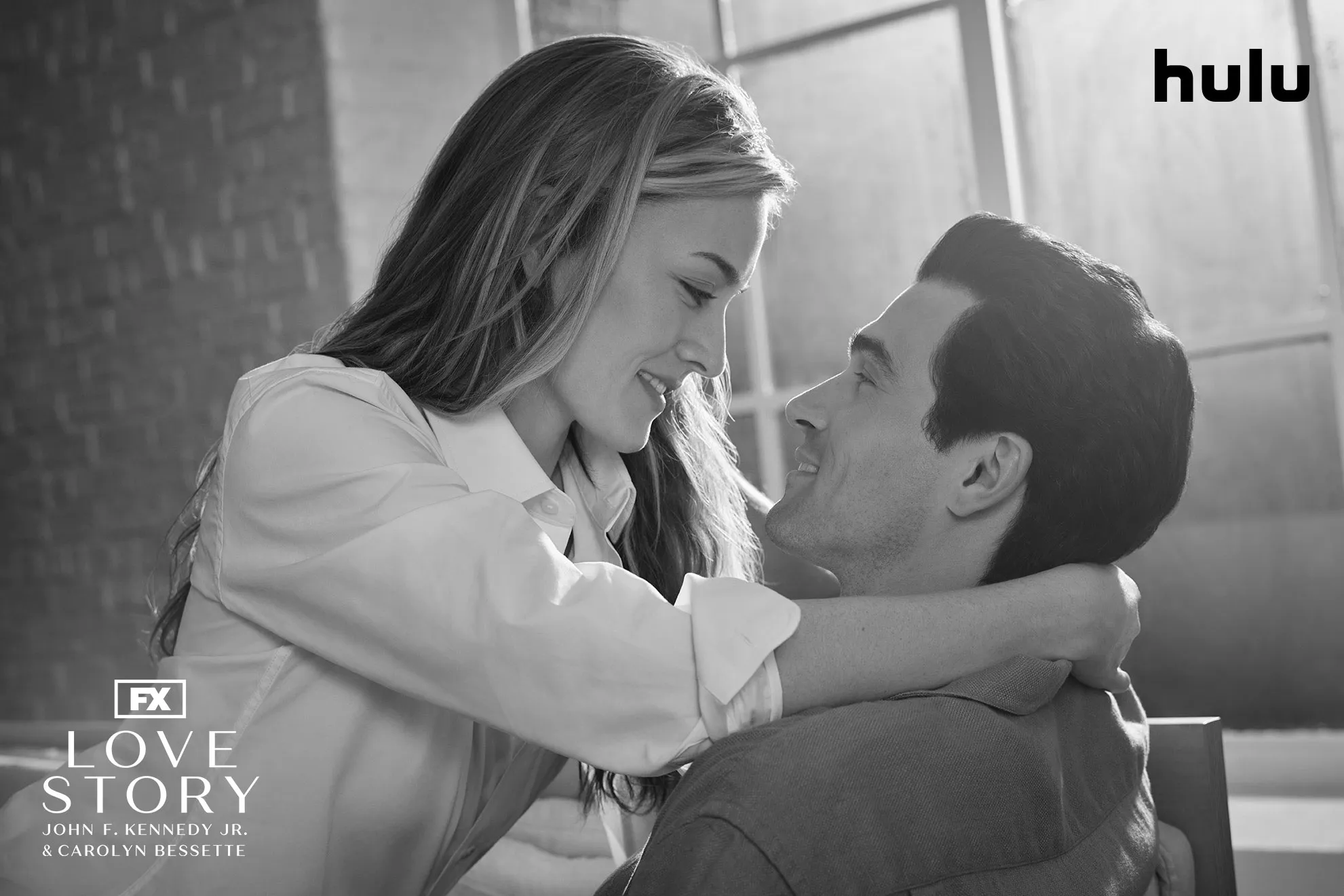 Carolyn Bessette (Sarah Pidgeon) and John F. Kennedy Jr. (Paul Kelly) embrace in a black and white set photo from FX's Love Story.
