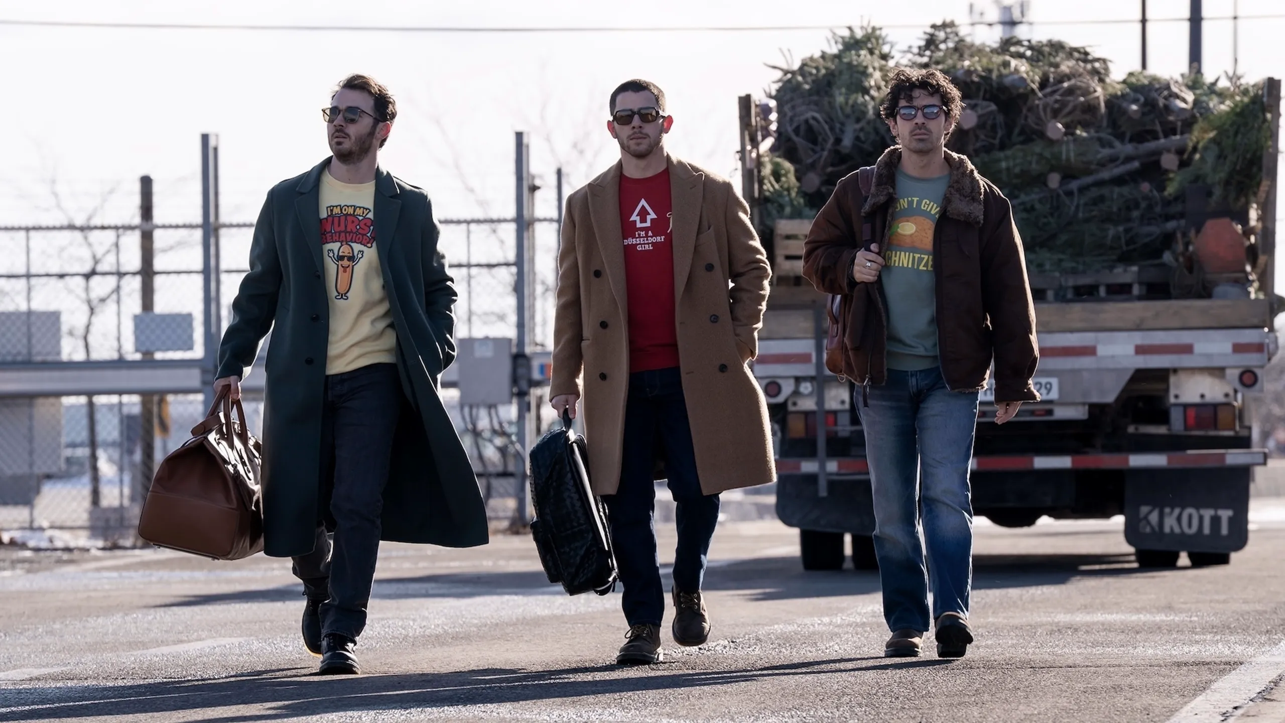 (L-R:) Kevin, Nick, and Joe Jonas walking along a road in winter carrying their bags.