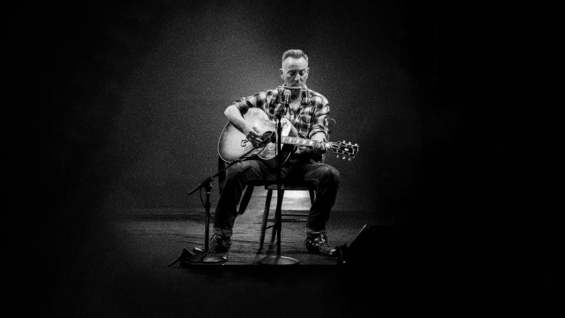 Black and white image of Bruce Spingsteen seated in the center of a stage playing a guitar during his tour.