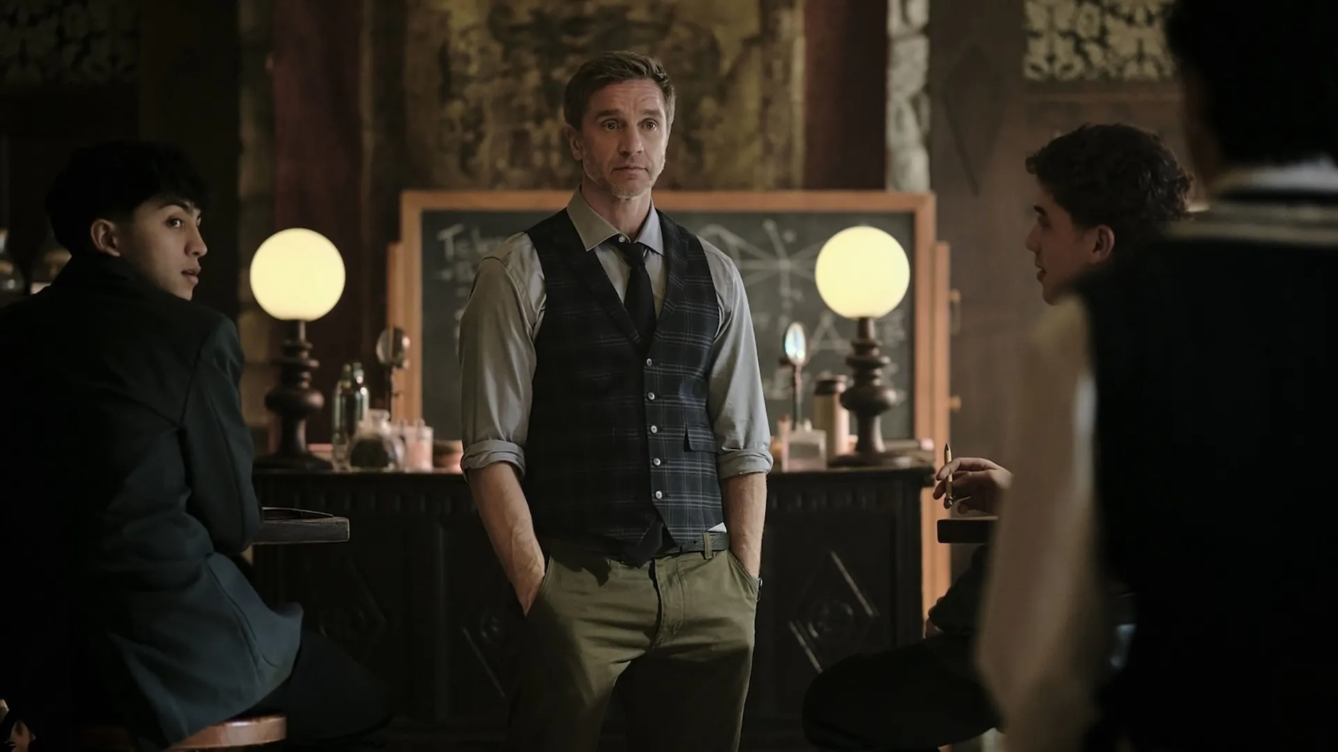 In a classroom scene from the supernatural series Coven Academy, Mr. Cole (Devon Sawa) stands with his hands in his pockets, wearing a plaid vest over a button-up shirt, while students look on from their desks.