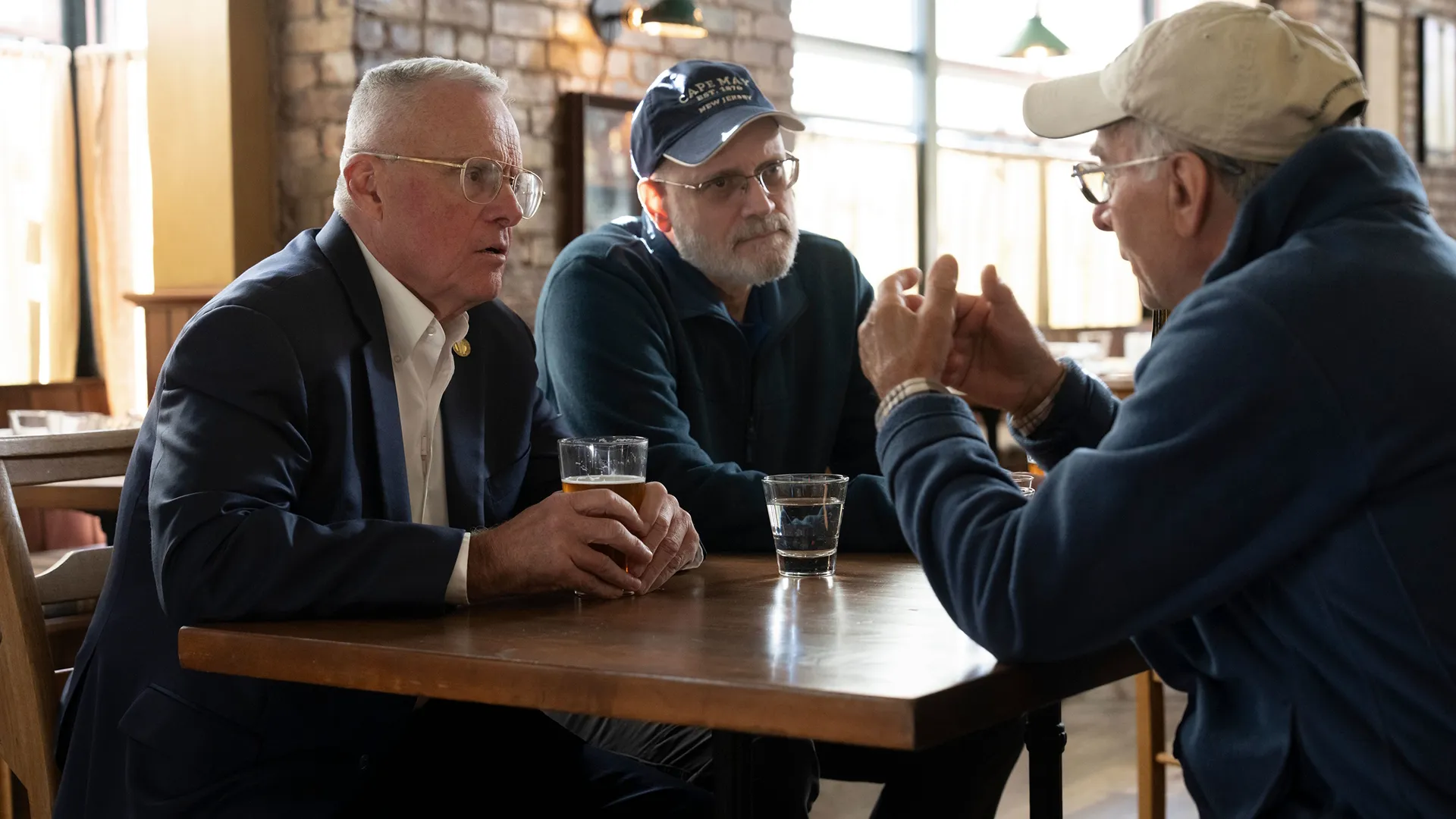 This image shows three men having a conversation at a wooden table in a pub setting, with one man in a suit wearing glasses and another in a cap.