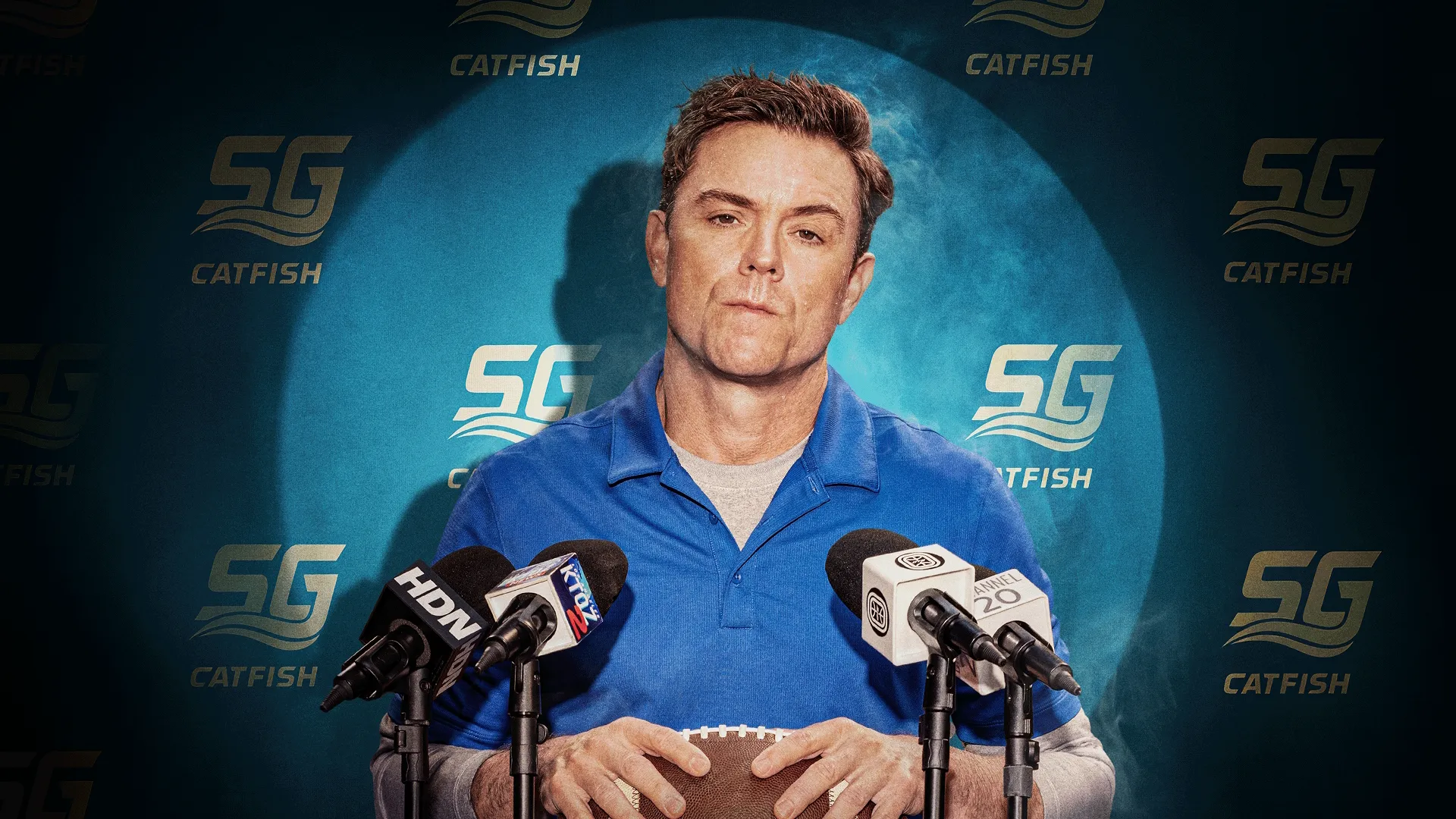 O treinador Scott Dobbs (Clayne Crawford) sentado com uma expressão cética atrás de uma fileira de microfones, segurando uma bola de futebol americano, posicionado em frente a um fundo azul com o logotipo do time "SG CATFISH".