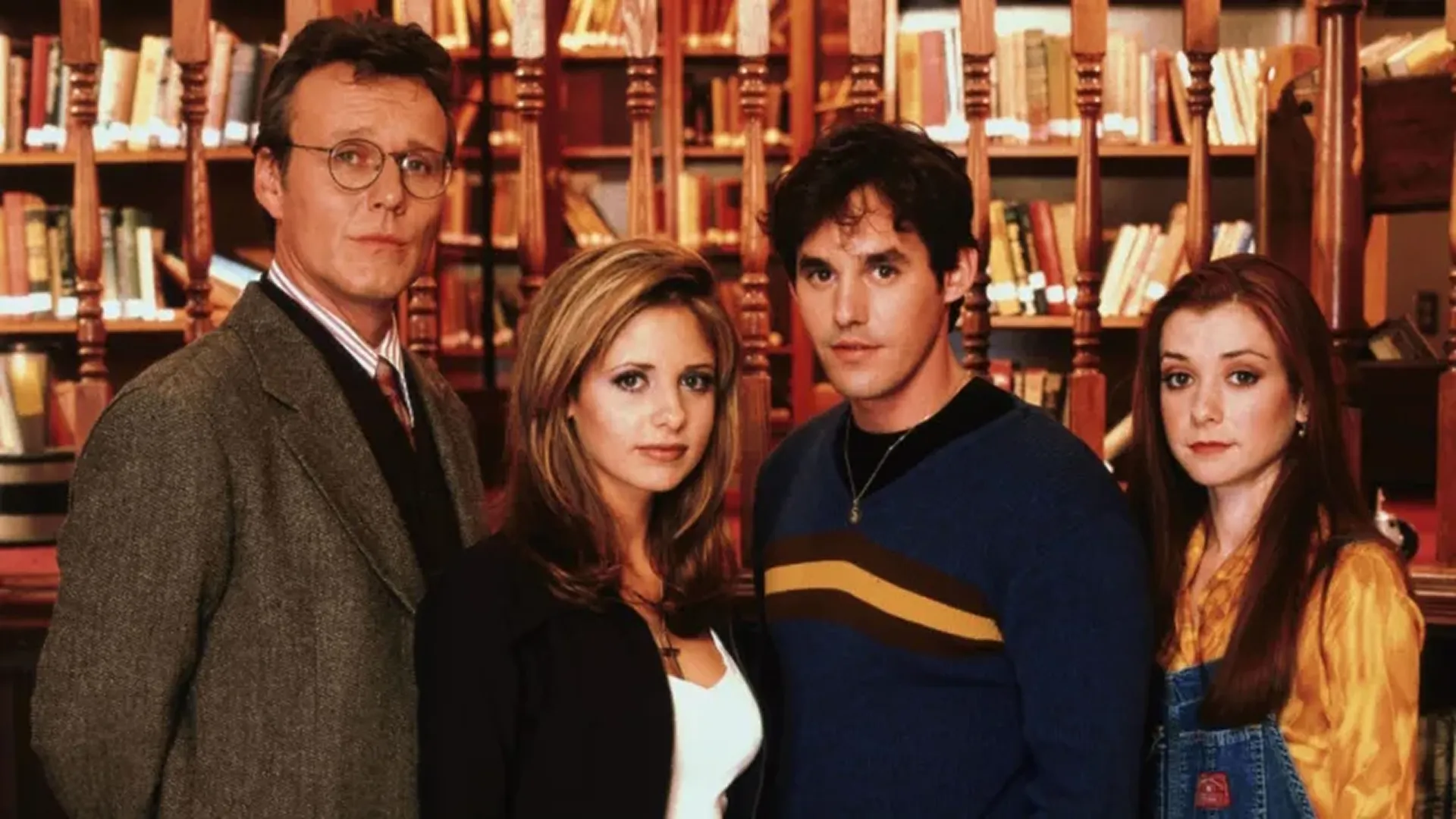 Rupert Giles (Anthony Head), Buffy (Sarah Michelle Gellar), Xander Harris (Nicholas Brendon), and Willow (Alyson Hannigan) looking towards the camera inside a library.
