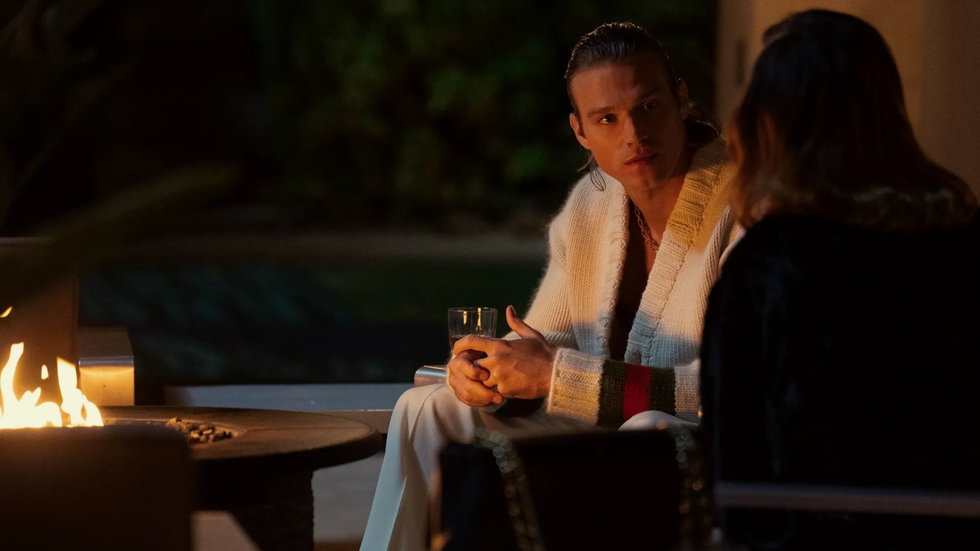 Chase Munroe (Matthew Nozska) sitting by a firepit at night talking to a woman.