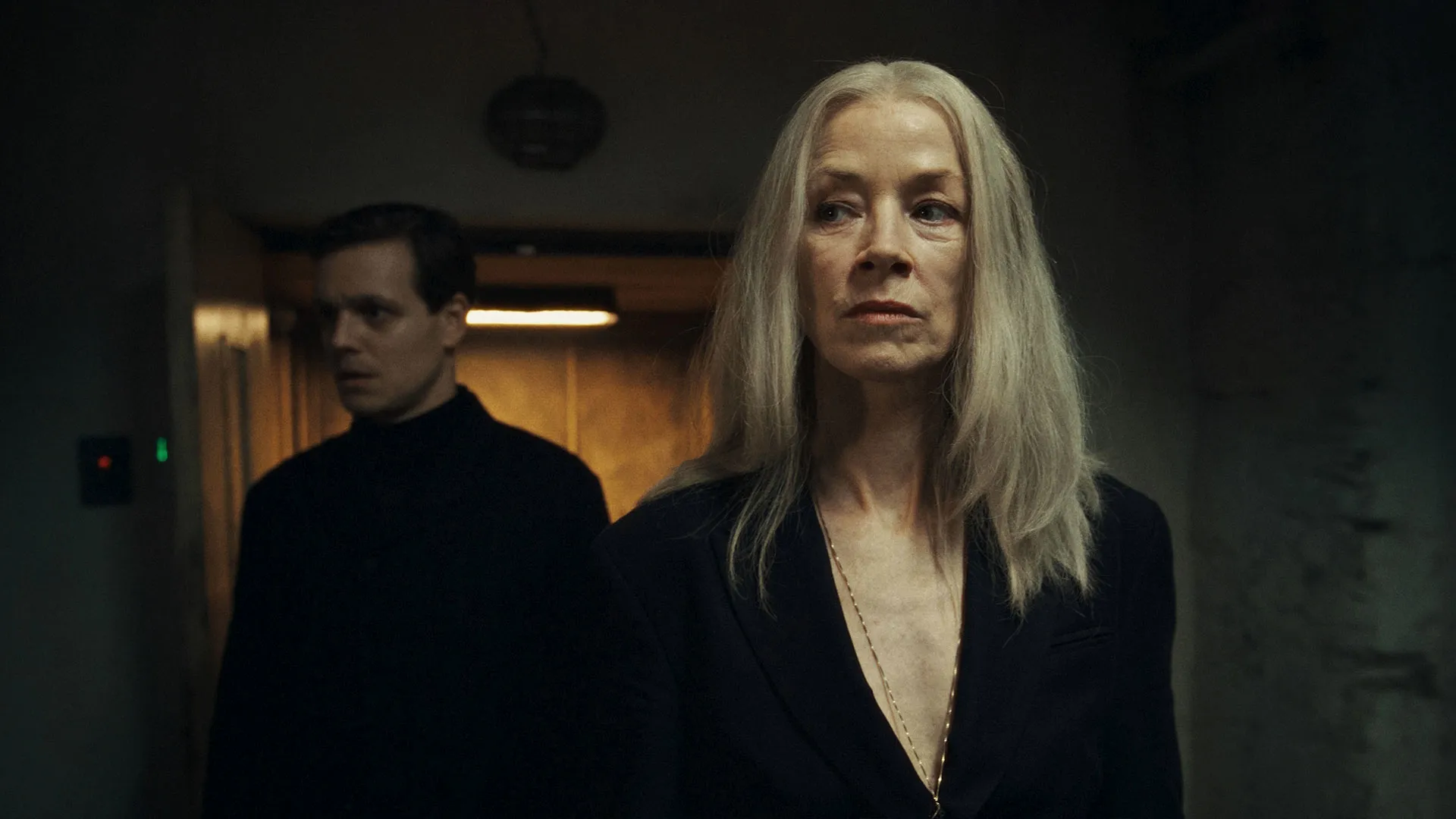 A person with long grey hair stands in the foreground while another person is visible in the background of a dimly lit hallway.