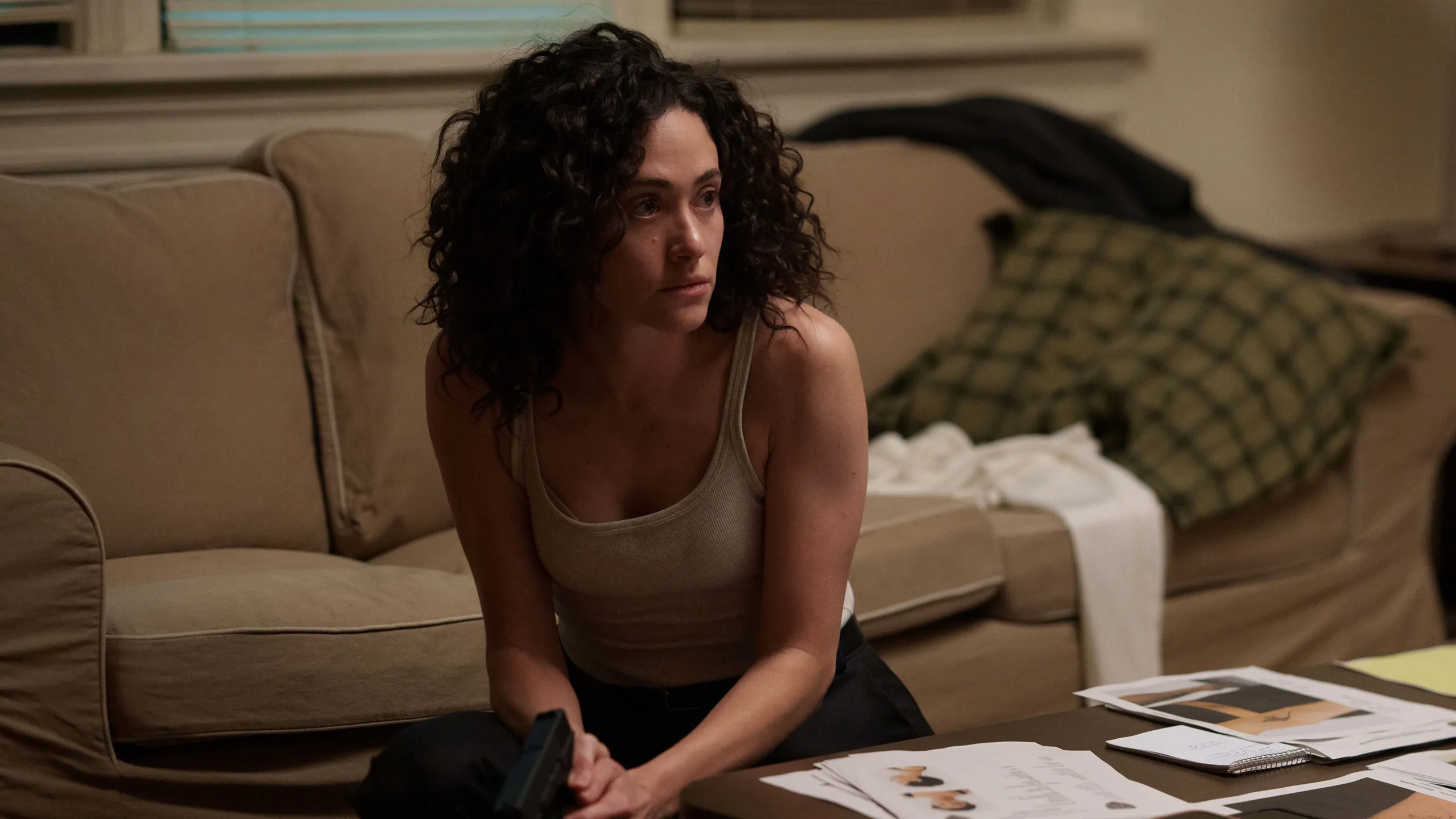 Alice crouches behind a coffee table surrounded by investigation files.