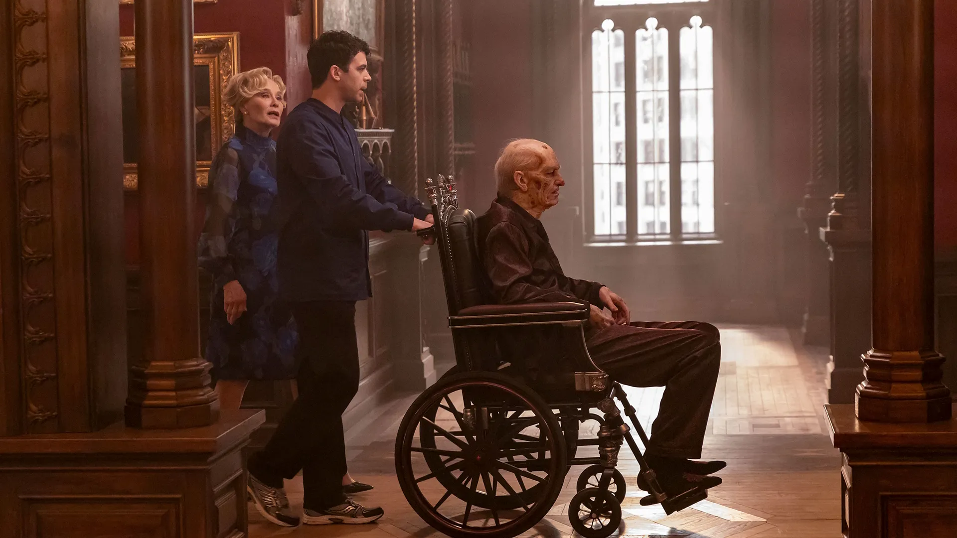 A young man pushes a frail elderly man in an ornate wheelchair down a dimly lit, grand hallway with large windows at the end, while a woman in a blue floral dress follows closely behind.