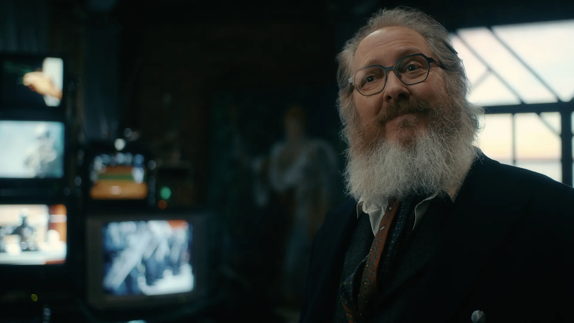 James Spader reprises his role as the human avatar form of Ultron, sporting a full gray beard, thick-rimmed glasses, a dark jacket, and a patterned tie, smiling subtly in front of stacked vintage television monitors in Marvel Television's VisionQuest.