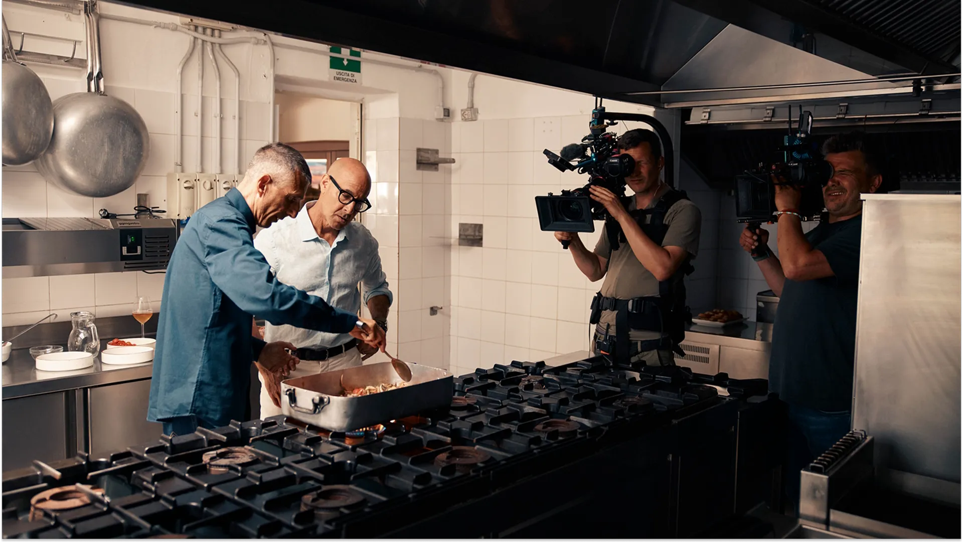 A camera crew films Stanley Tucci watching a chef cook in a restaurant kitchen