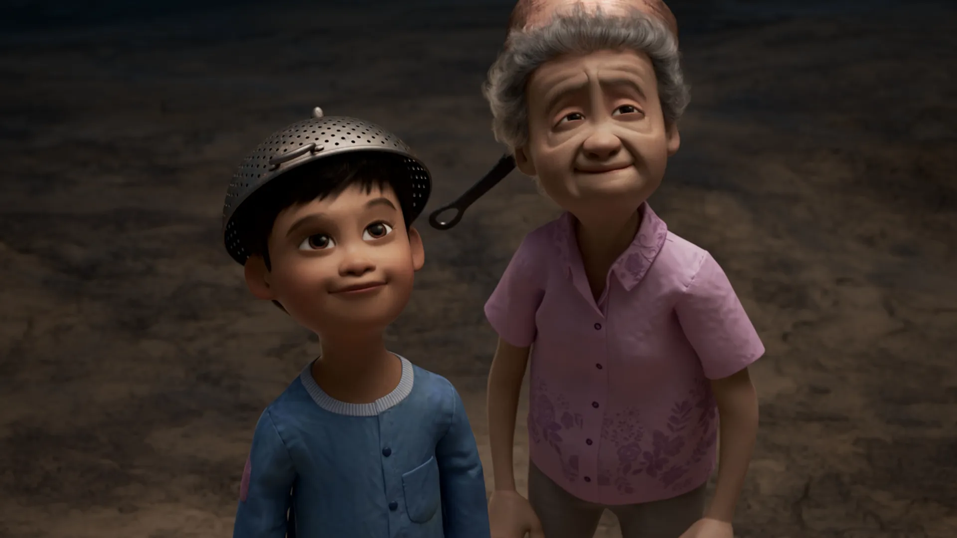 A young boy wearing a colander on his head smiles upwards with his grandmother, who is wearing a pan on her head