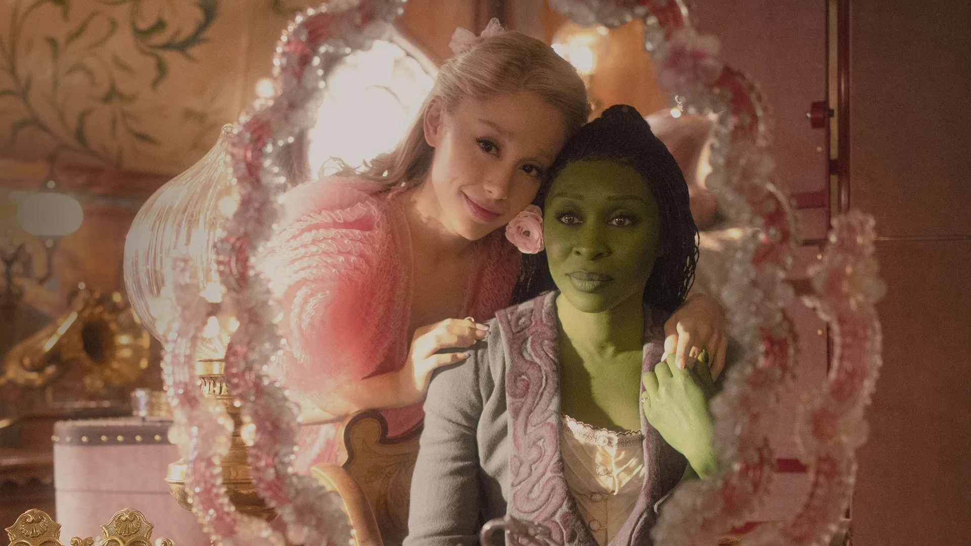 Ariana Grande as Glinda smiles softly, resting her head against Cynthia Erivo as Elphaba, who is green-skinned and looking forward, as they are reflected together in an ornate, pink-framed vanity mirror in a scene from the film Wicked.