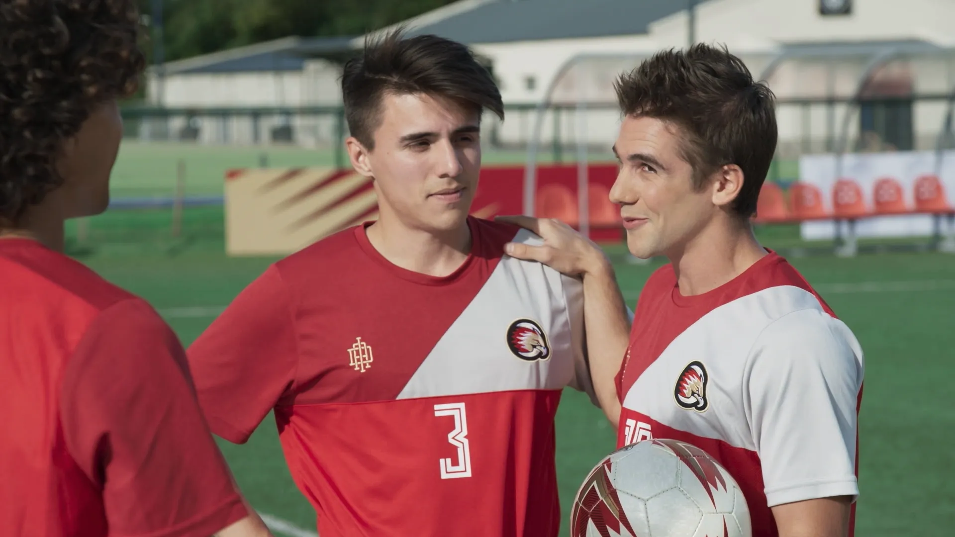 A scene from the teen sports series O11CE showing soccer players Leo Palacios (Juan Baudilio) in jersey number 3 and Gabo Moreti (Mariano González-Guerineau) holding a soccer ball on a practice field as a teammate speaks to them.