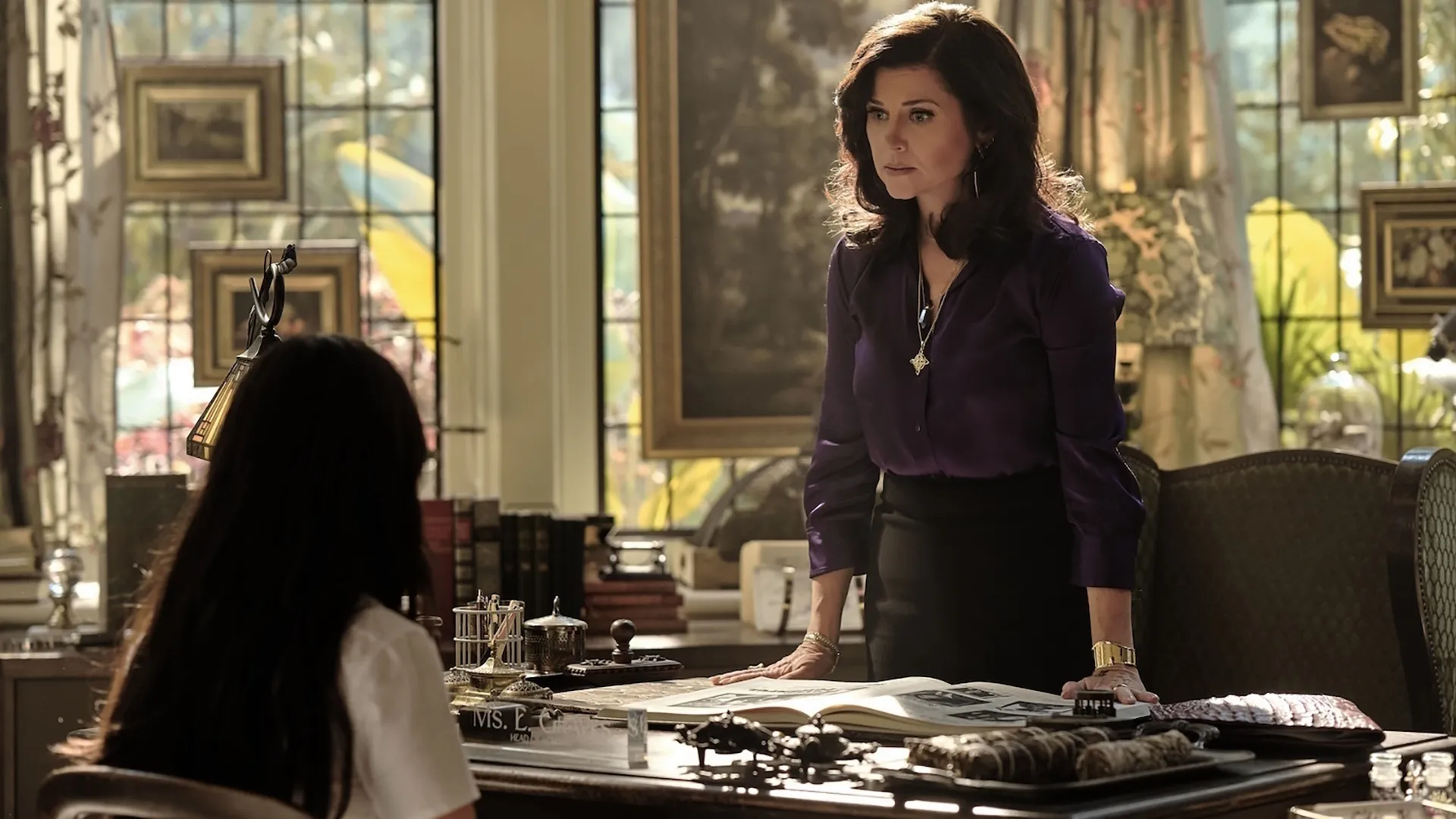 In an office scene from the supernatural series Coven Academy, Miss Graves (Tiffani Thiessen) stands leaning over a large desk, wearing a purple silk blouse and a gold cuff bracelet, as she looks strictly at a student sitting across from her.