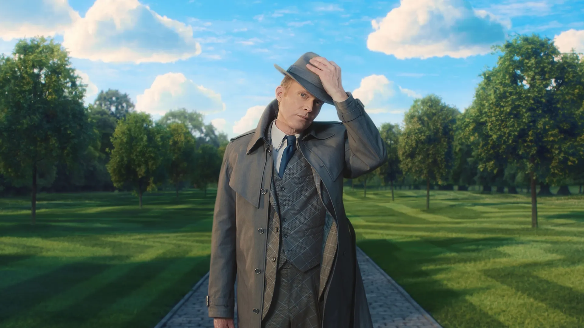 Vision in human disguise tipping his gray fedora while wearing a trench coat and a plaid three-piece suit, standing on a cobblestone path centered within a surreal, manicured park under a bright blue sky with puffy white clouds in Marvel Television's VisionQuest.