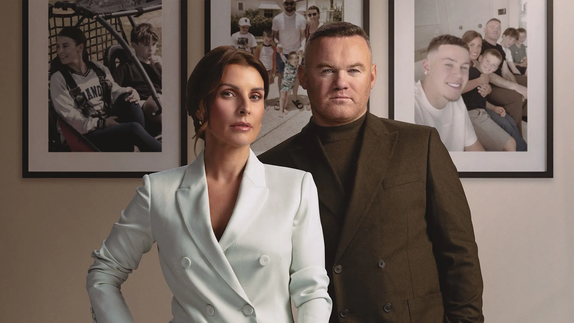 Wayne Rooney and Coleen Rooney pose together in front of a wall with three family portraits hanging on it