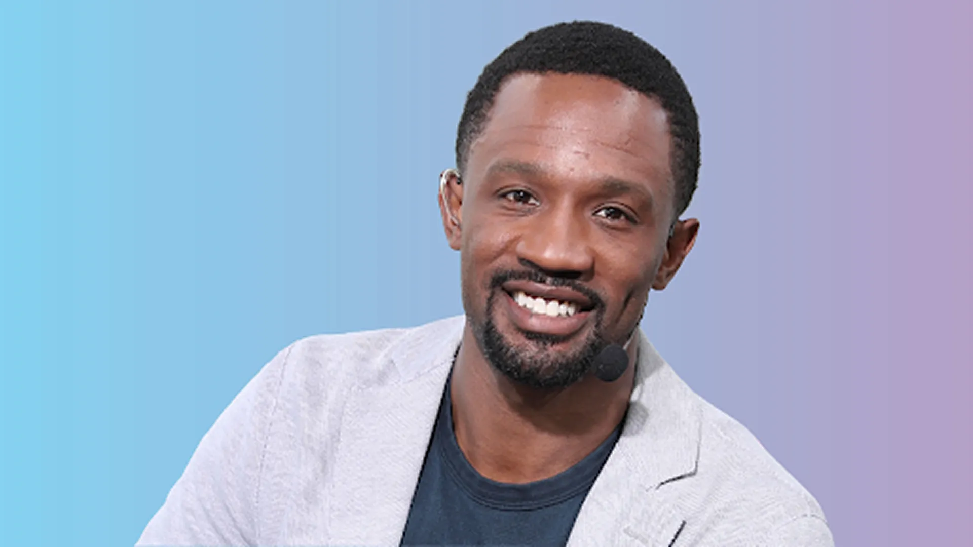 Promotional graphic for The Domonique Foxworth Show featuring host Domonique Foxworth smiling while wearing a light grey blazer, headset microphone, and earpiece against a blue-to-purple gradient background.
