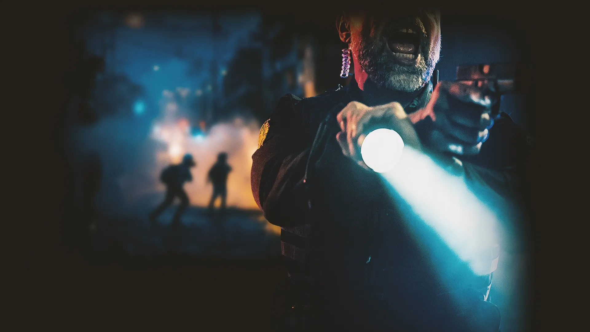 A police officer points a handgun and a bright flashlight forward while shouting, framed against a smoky, chaotic night street scene with silhouettes of running figures in the background.