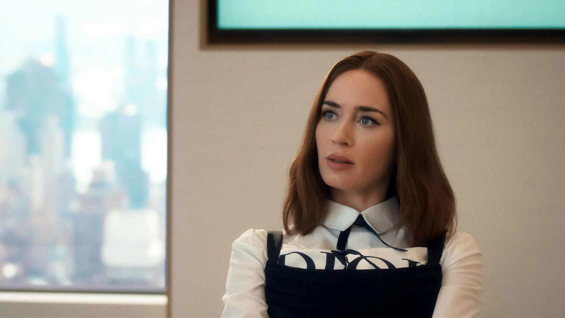 A promotional still from The Devil Wears Prada 2 shows Emily Charlton (Emily Blunt) looking off-camera with a serious expression inside a modern office. She has shoulder-length reddish-brown hair and wears a sharp white collared blouse layered underneath a black Christian Dior structural bodice.