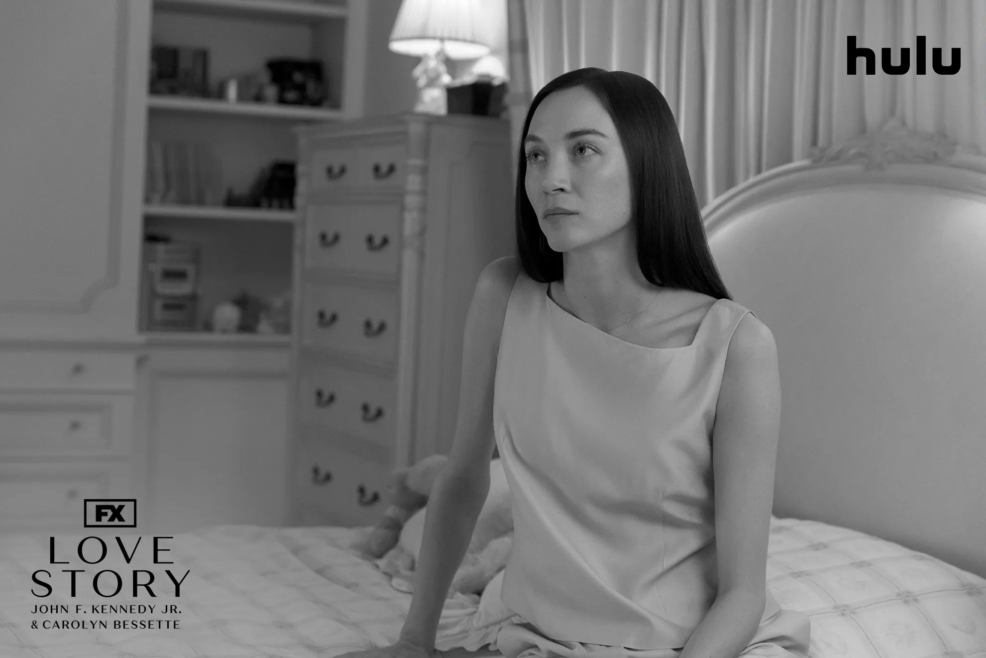 A brunette Carolyn Bessette (Sarah Pidgeon) sitting on a bed in a black and white set photo from FX's Love Story.