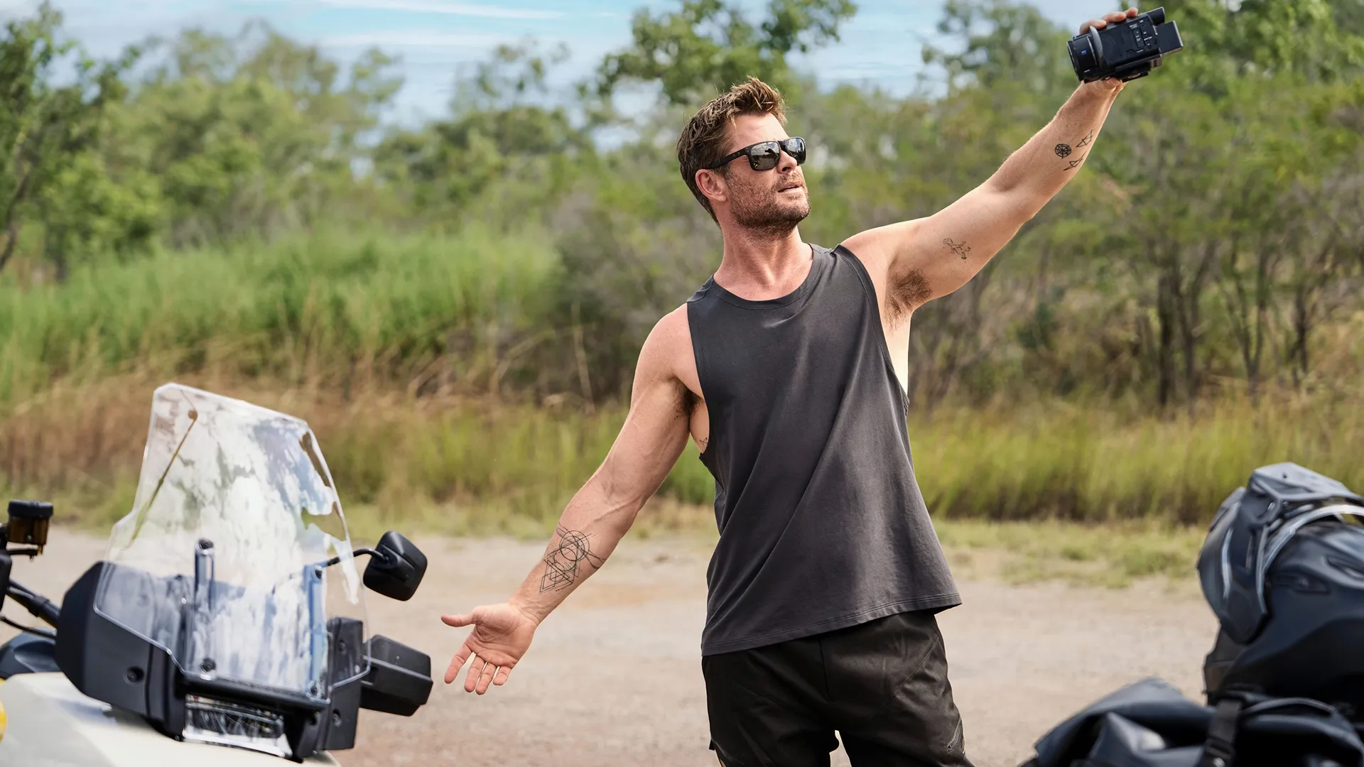 Chris Hemsworth looking up at the sky while wearing sunglasses in the Austarlian wilderness. Standing next to two motorcycles.