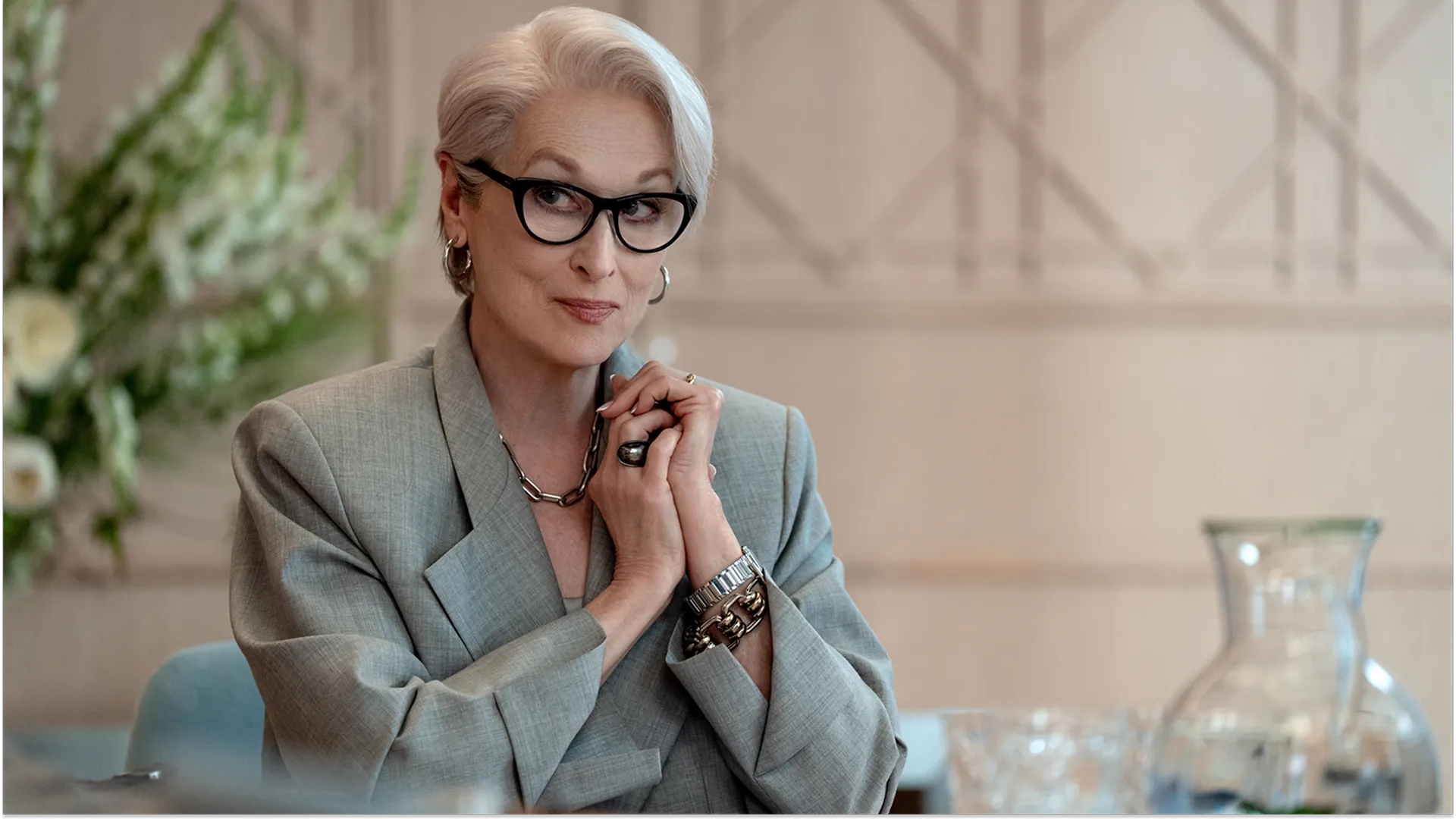 A promotional still from The Devil Wears Prada 2 shows Miranda Priestly (Meryl Streep) seated at a table, looking forward with a knowing smile and her hands clasped together. She sports her signature styled white hair, thick black-framed glasses, silver hoop earrings, a thick chain necklace, and a tailored grey oversized blazer.