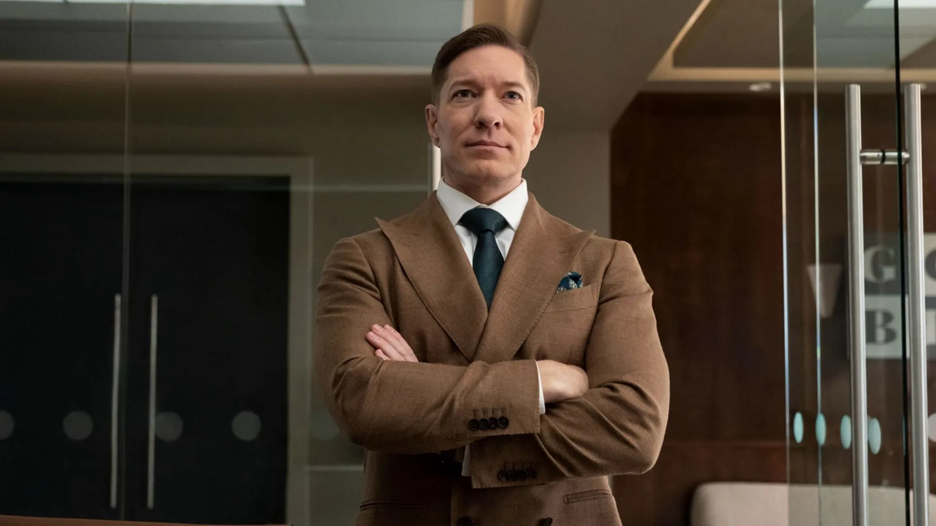 Joseph Sikora as Bill Sterling, wearing a brown suit, standing with his arms crossed