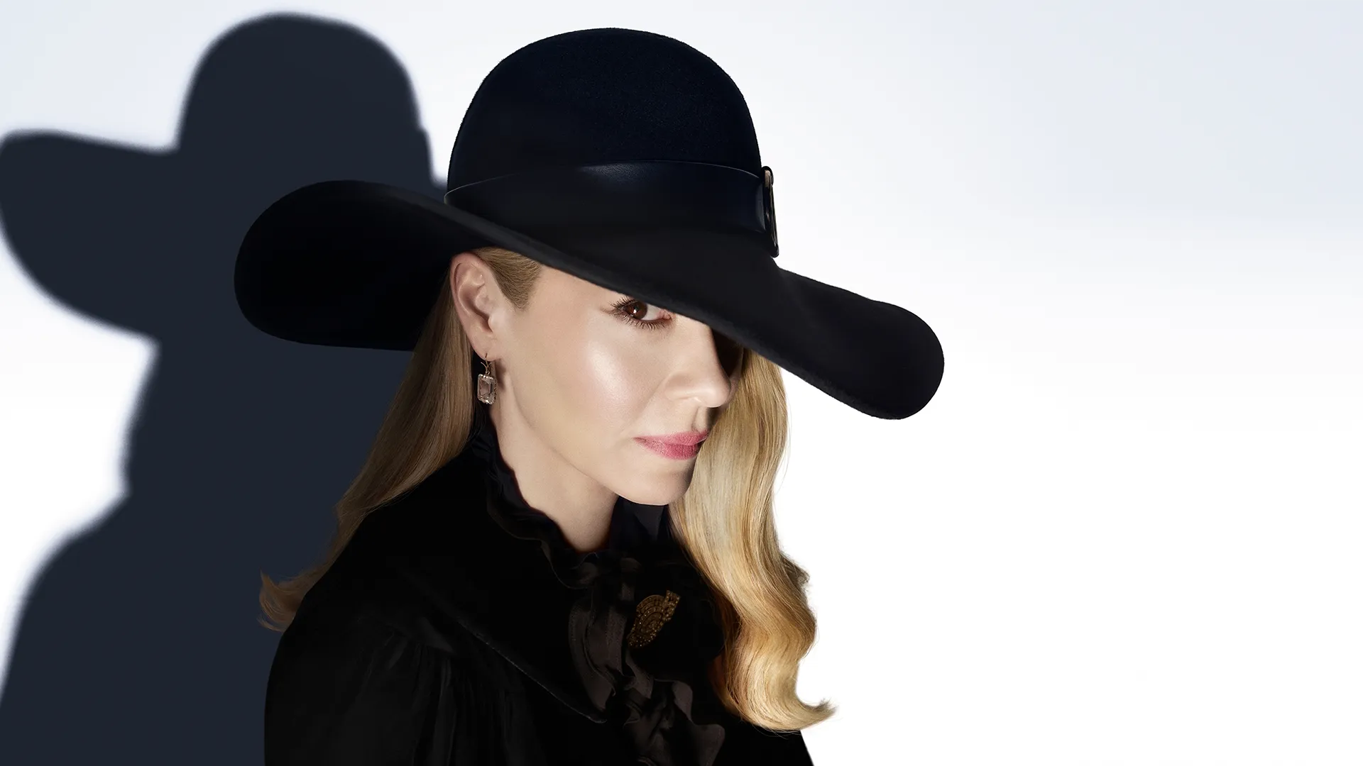 A blonde woman in profile wears a wide-brimmed black hat and matching high-collared shirt, casting a sharp shadow against a bright white background.