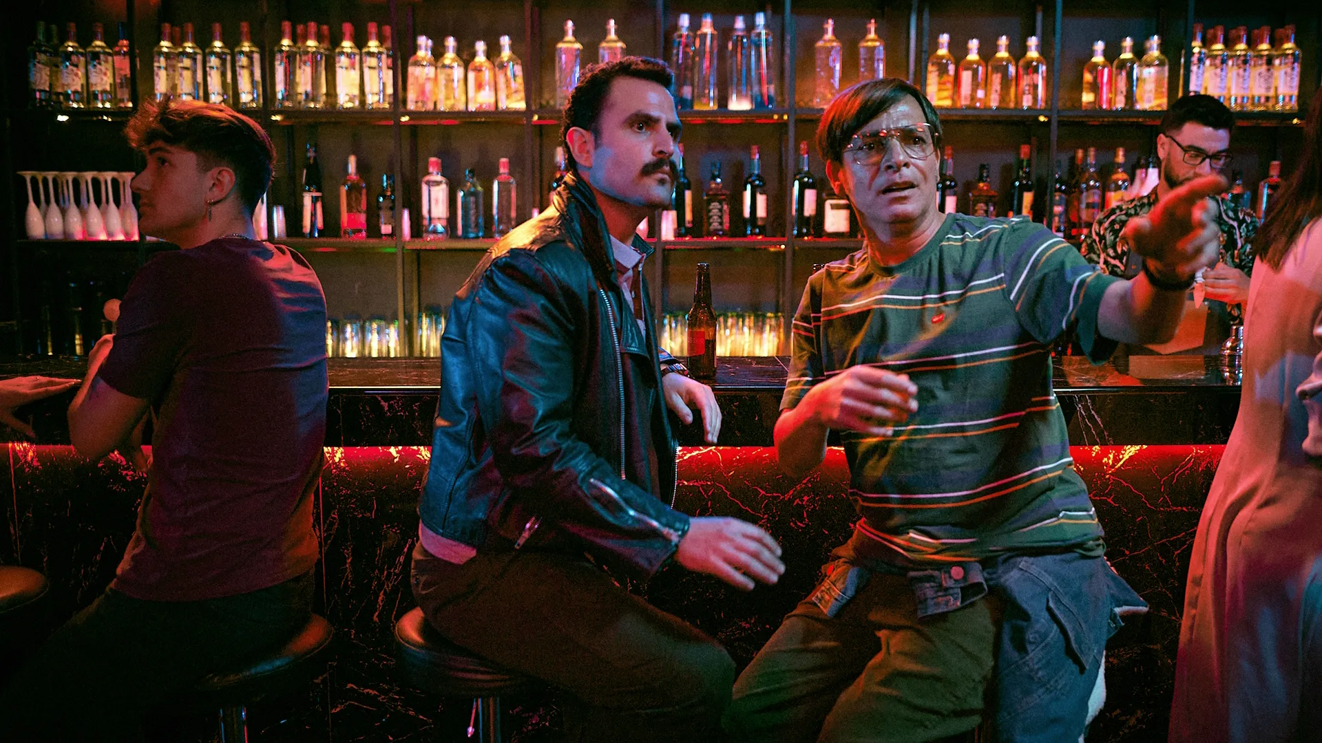 An official promotional image from the Argentine comedy-drama series Amores inesperados, featuring two men sitting at a dimly lit bar, with one man pointing forward into the distance.