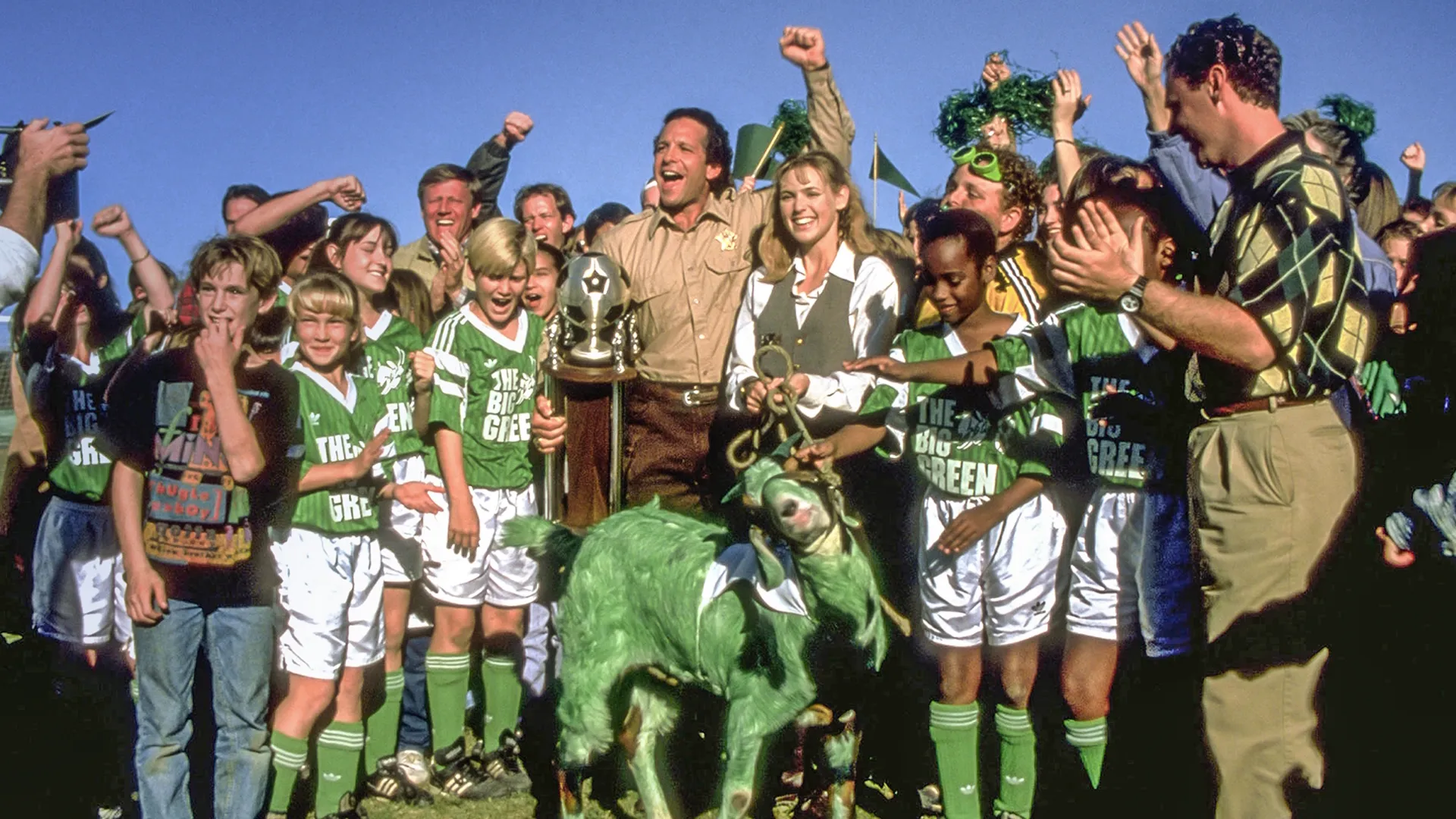 A celebratory scene from the sports comedy film The Big Green showing Sheriff Tom Palmer (Steve Guttenberg) and Miss Anna Montgomery (Olivia d'Abo) cheering with a gold trophy alongside their youth soccer team, who are gathered around a live goat painted completely green.
