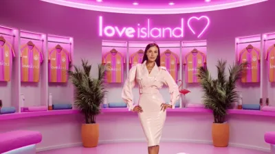 Maya Jama stands in a pink, neon-lit Love Island locker room wearing a latex trench dress, surrounded by hanging sports jerseys.