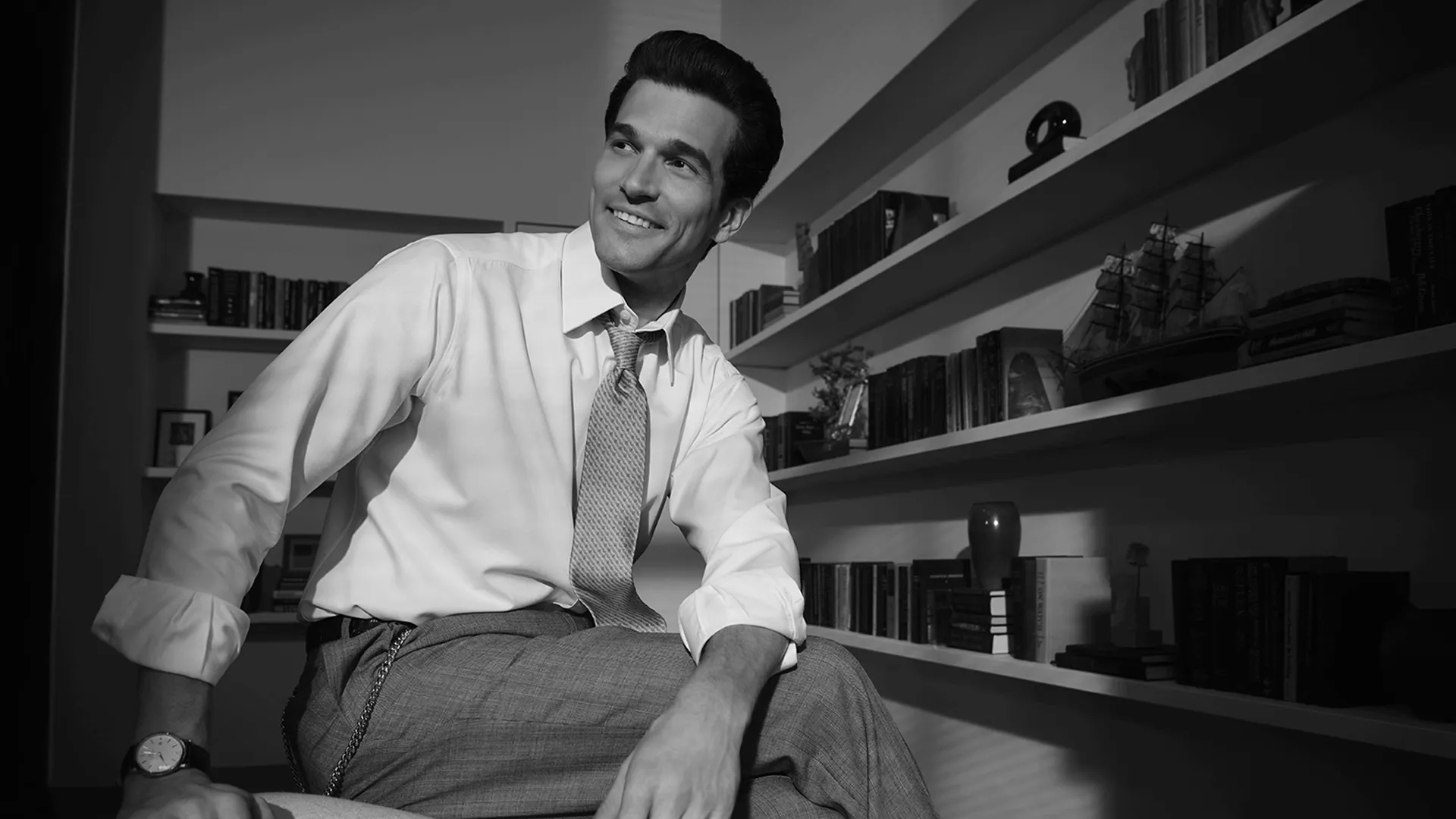 A smiling John F. Kennedy Jr. (Paul Kelly) sitting in an office in a black and white set photo from FX's Love Story.