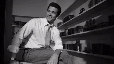 Foto em preto e branco de John F. Kennedy Jr. (Paul Kelly) sorrindo, sentado em um escritório, em História de Amor, da FX.