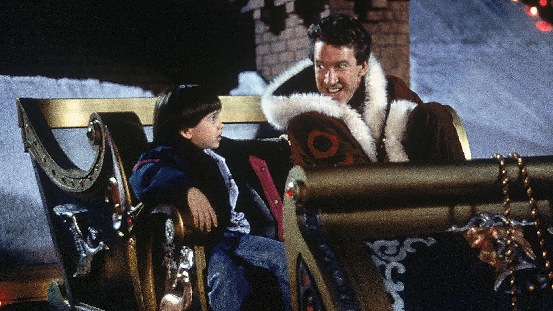 Scott Calvin (Tim Allen), dressed in a Santa suit, and Charlie Calvin (Eric Lloyd) sit in Santa's sleigh on a snow-covered rooftop.