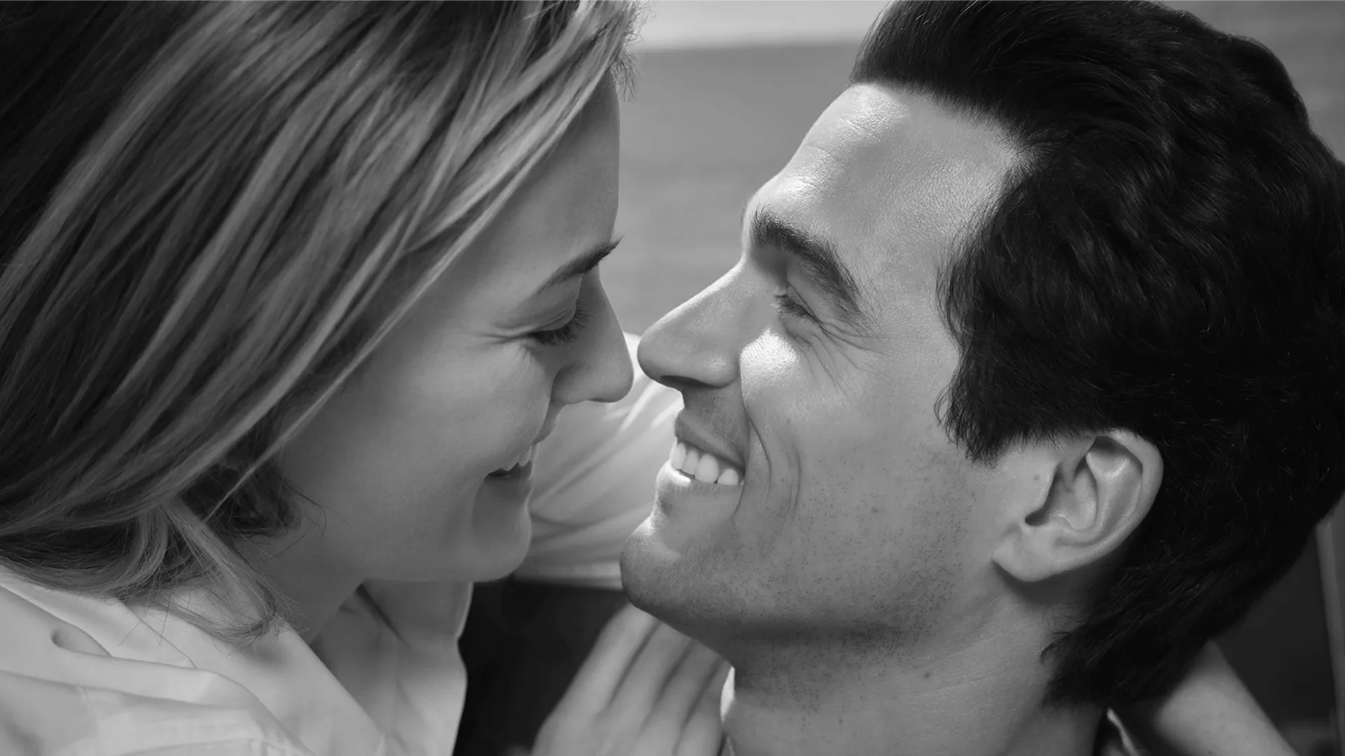 Close-up of Carolyn Bessette (Sarah Pidgeon) and John F. Kennedy Jr. (Paul Kelly) embracing in a black and white set photo from FX's Love Story.