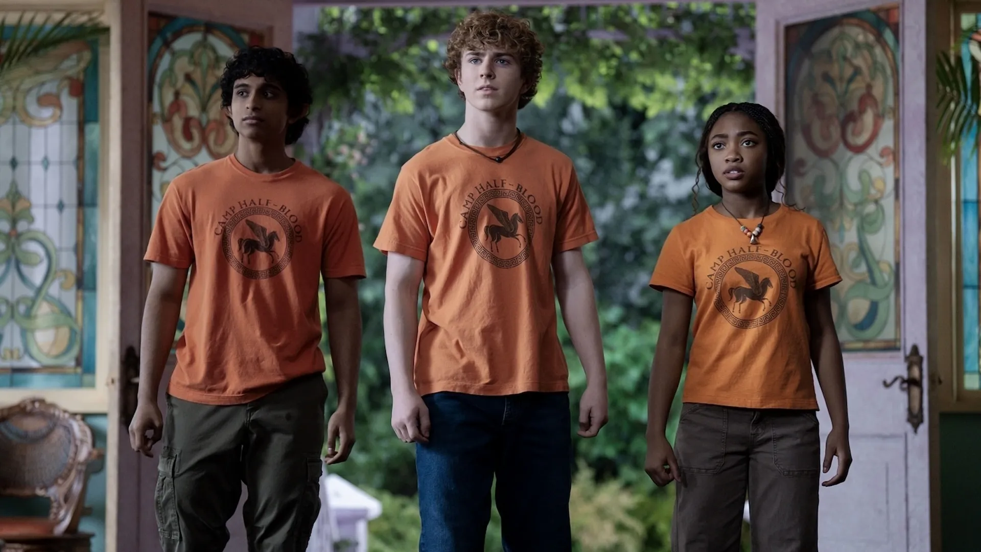 Grover Underwood (Aryan Simhadri), Percy Jackson (Walker Scobell), and Annabeth Chase (Leah Sava Jeffries) stand side-by-side in matching orange Camp Half-Blood t-shirts, looking forward with tense, determined expressions in front of a wide doorway.