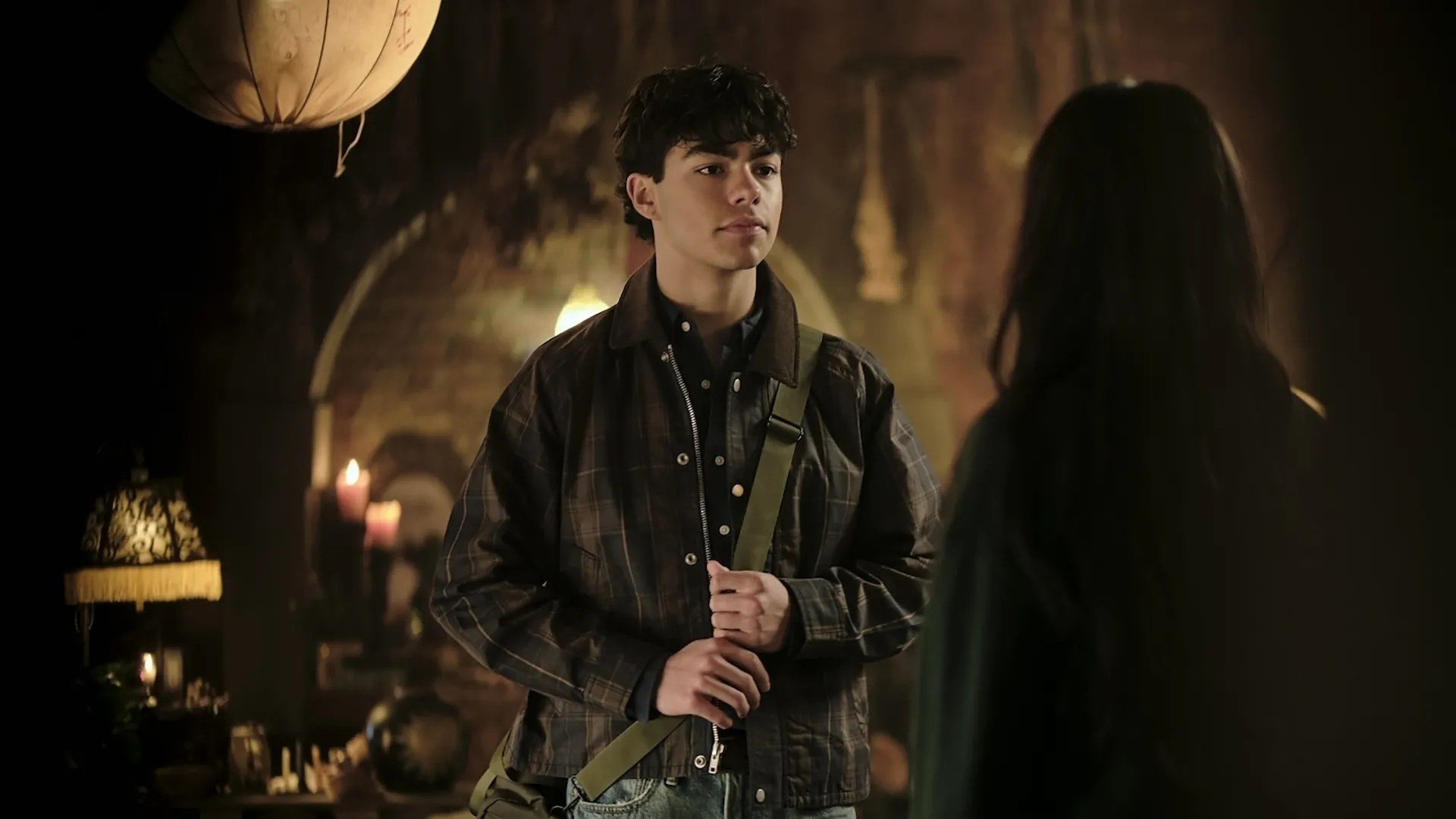 In a scene from the supernatural series Coven Academy, Jake (Malachi Barton) stands in a dimly lit, mystically decorated room wearing a plaid jacket and a green shoulder bag while speaking to a girl whose back is turned to the camera.