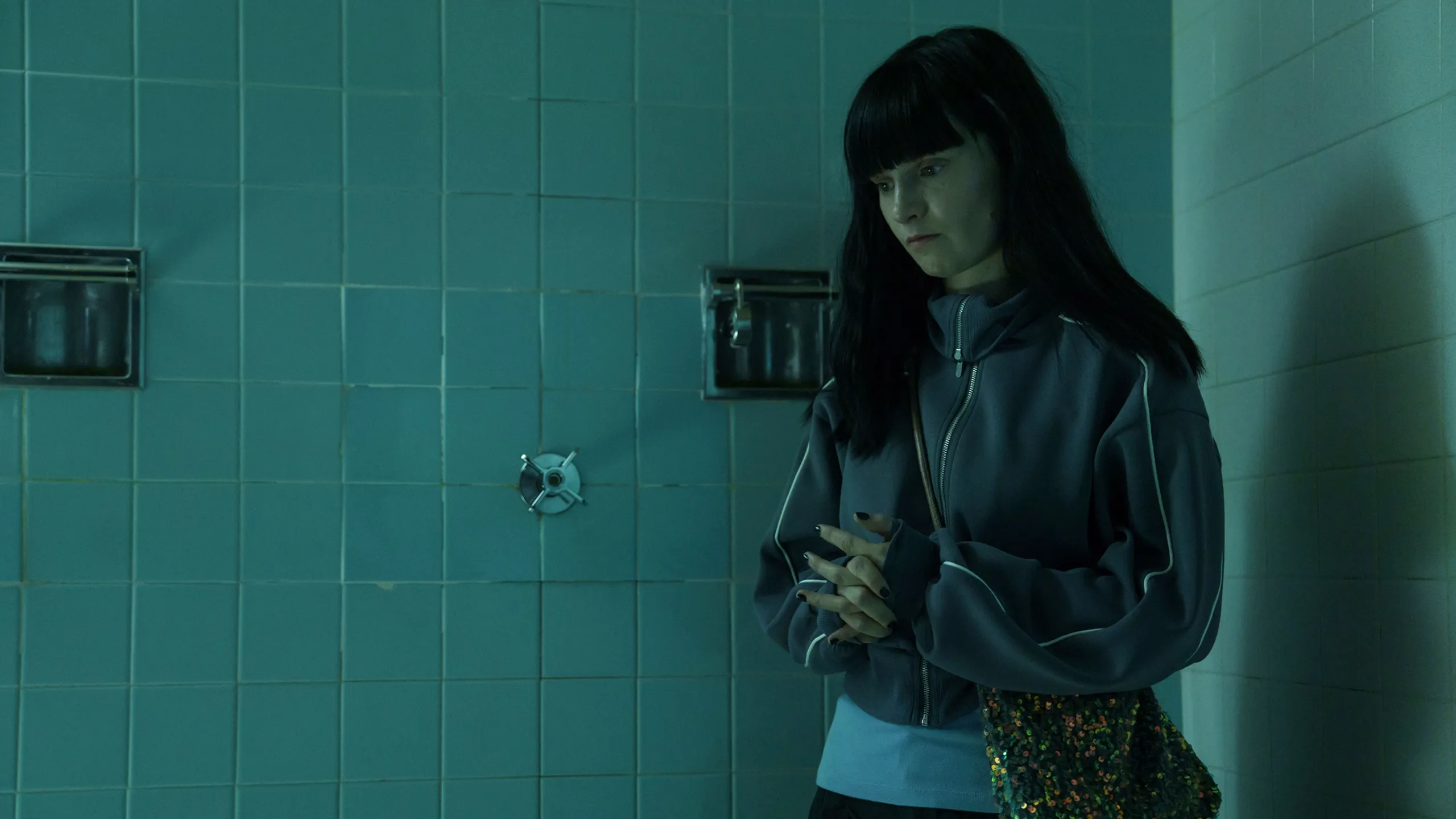 Catherine stands in a tiled bathroom carrying a sequined bag.
