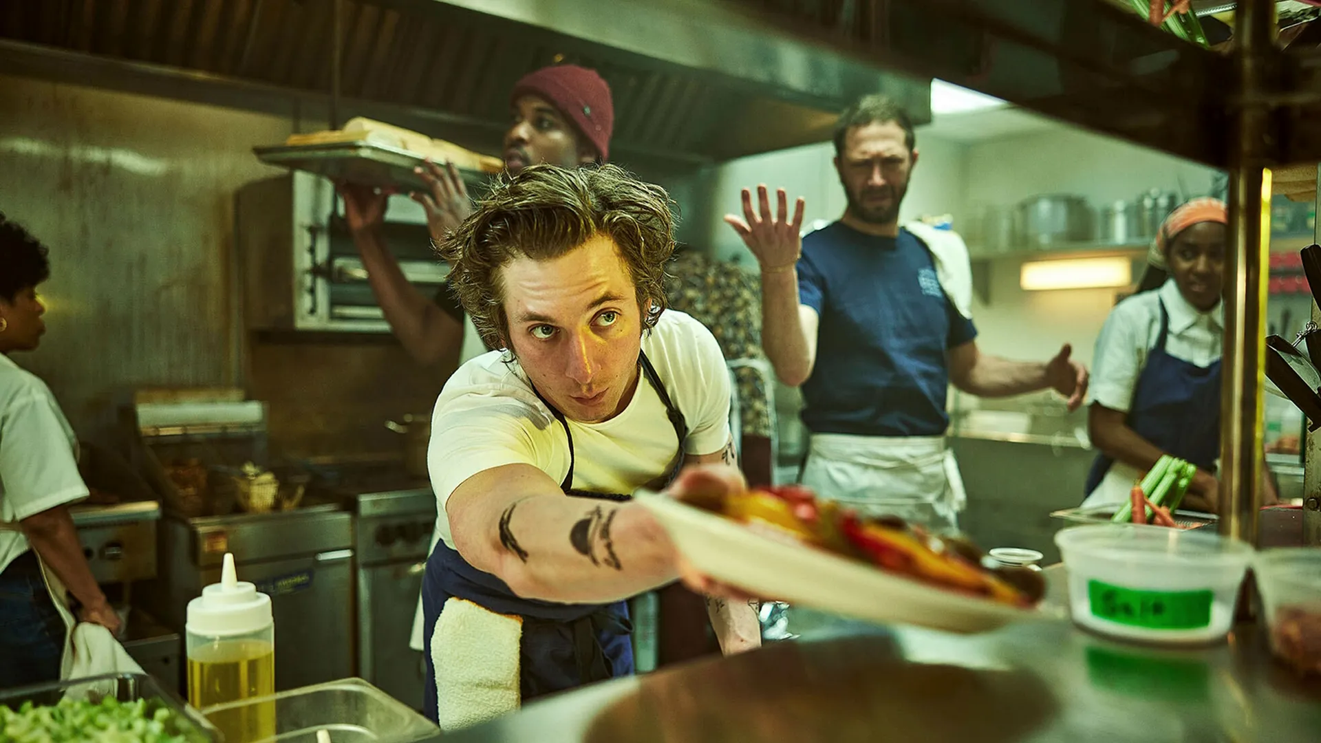 Carmy handing a plate through the pass of a kitchen, with other people working in the background