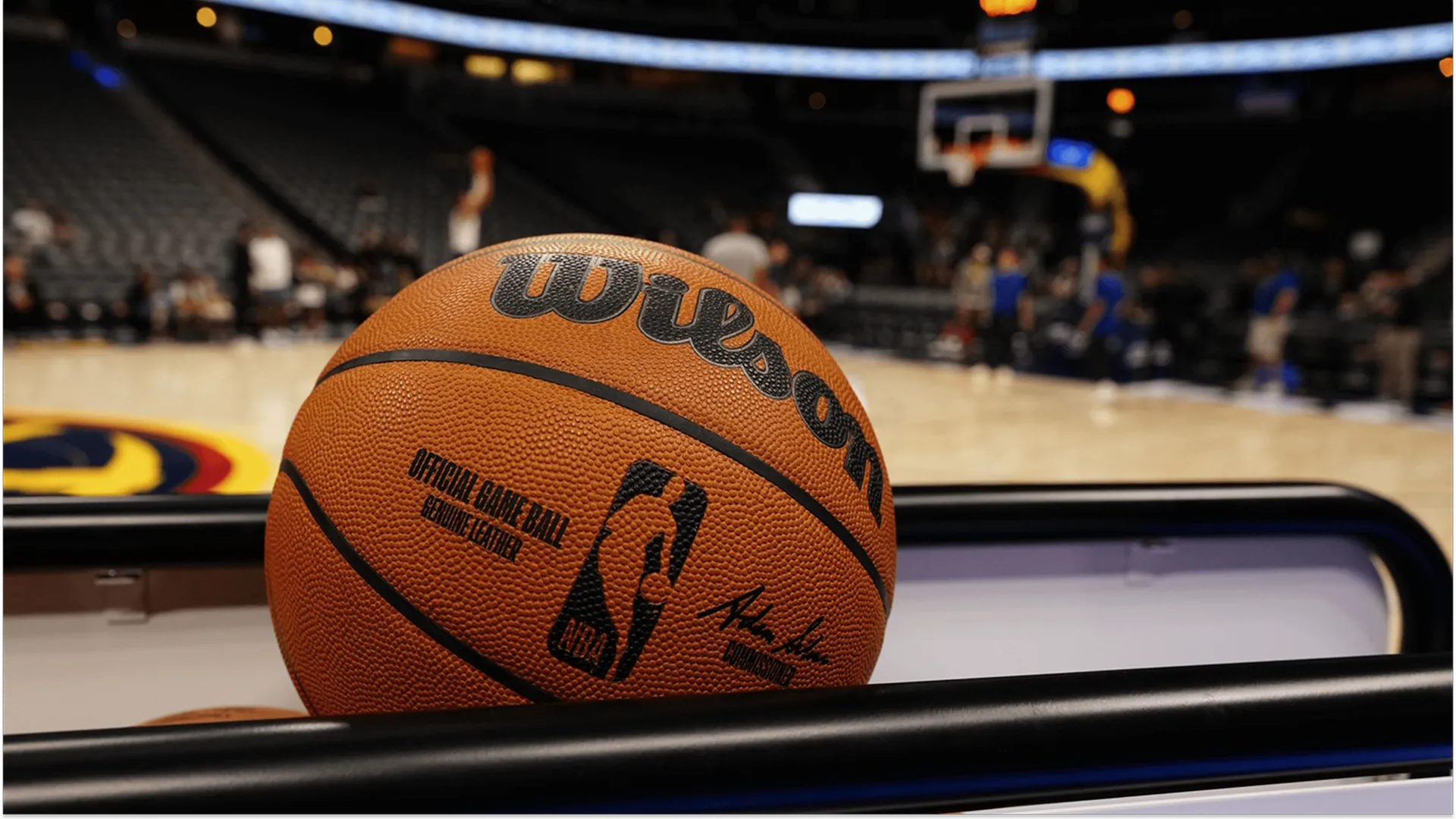 A close-up shot of an official Wilson NBA basketball resting on a rack, with a blurred basketball court and arena seats in the background.