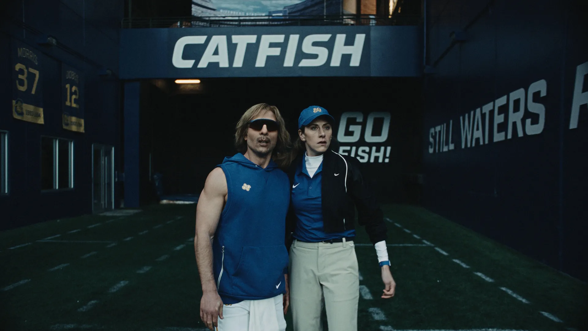 Chad Powers (Glen Powell) e Ricky (Perry Mattfeld) em um campo de futebol americano, em frente a uma parede do estádio que diz "CATFISH GO FISH!", vestidos com roupas esportivas azuis do mesmo time.