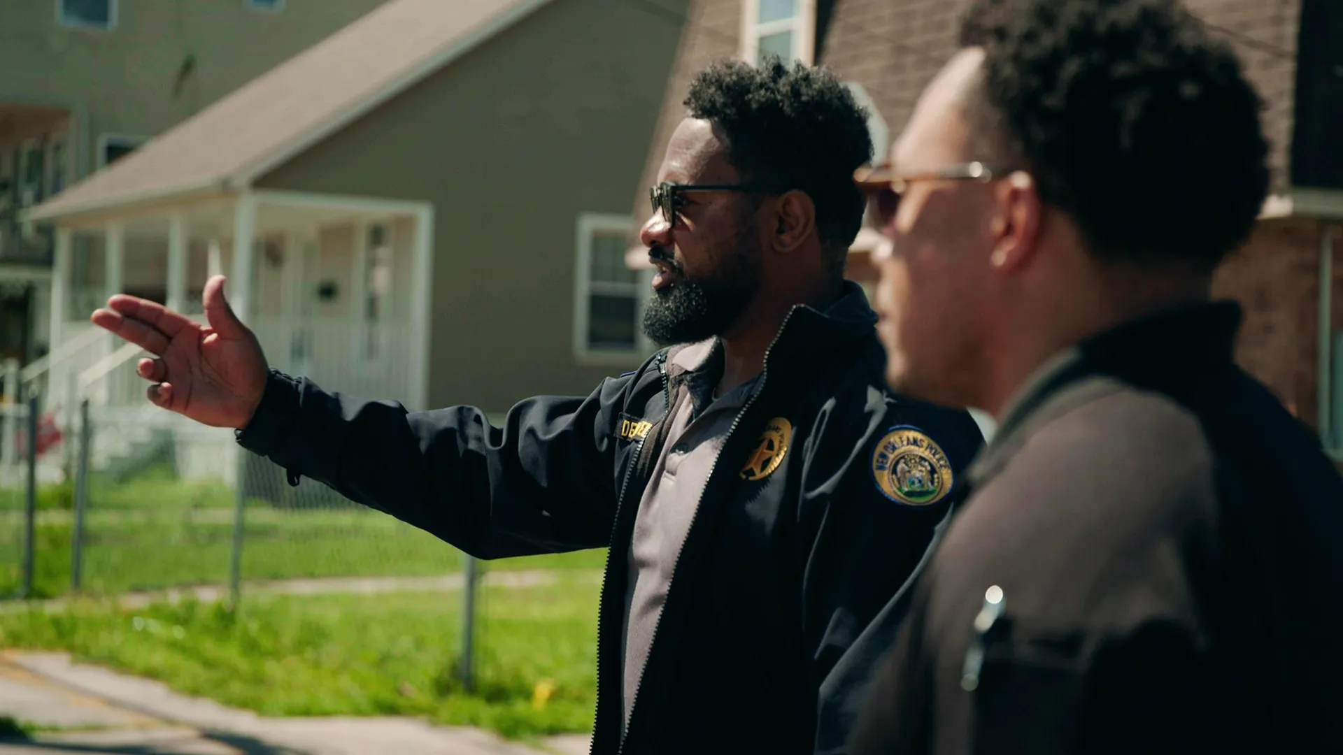 New Orleans Police Department detective Stewart stands outdoors wearing sunglasses and a black uniform jacket, extending his arm to point across a residential street while talking with a colleague in a scene from the true crime docuseries Homicide Squad: New Orleans