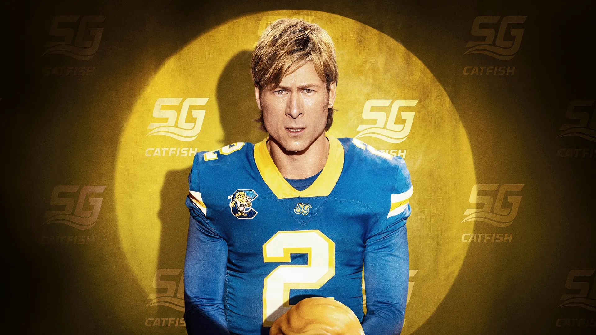 Chad Powers (Glen Powell) olha intensamente para frente com uma expressão concentrada, vestindo uma camisa de futebol americano do "SG CATFISH" azul e amarela, com o número 2, em frente a um fundo amarelo brilhante iluminado por holofotes.