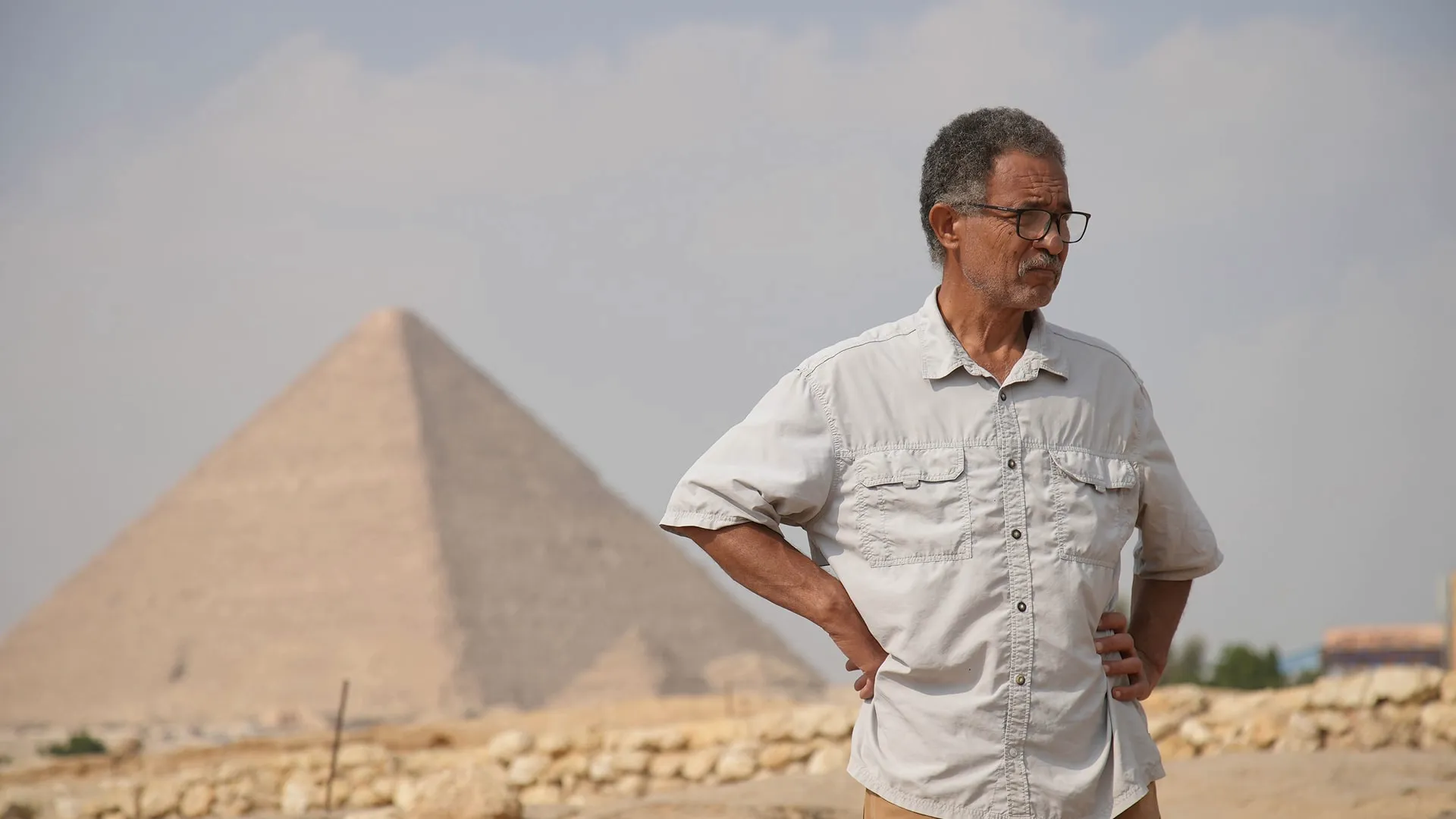 Archaeologist wearing a white shirt and glasses standing with his hands on his hips with a pyramid looming in the background.