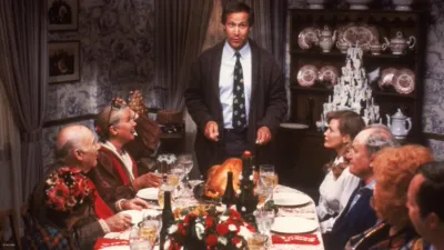 Clark Griswold (Chevy Chase) stands at the head of a dining table decorated for Christmas, preparing to carve a turkey.