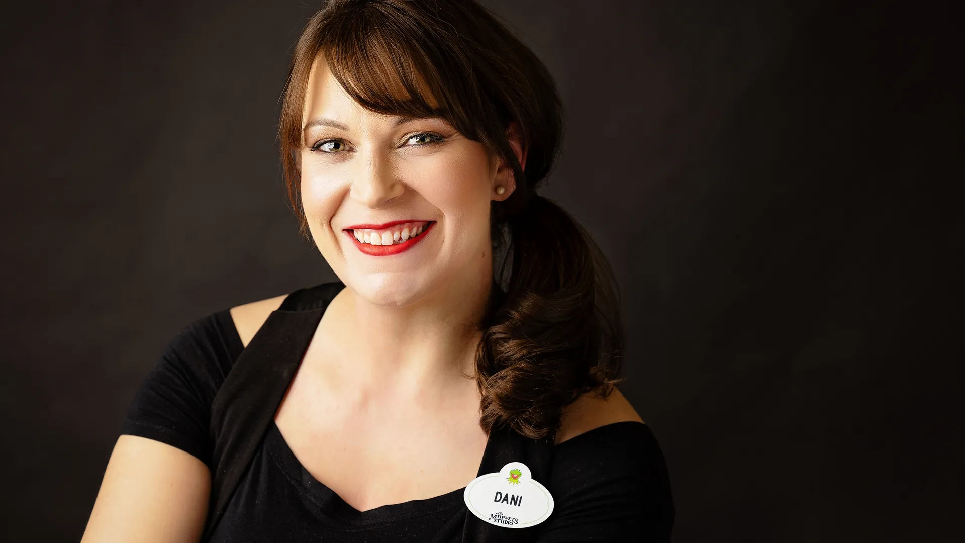 A headshot of Dani Iglesias, Creative Director of The Muppets Studio, smiling in front of a dark background and wearing a Muppets Studio employee name tag.